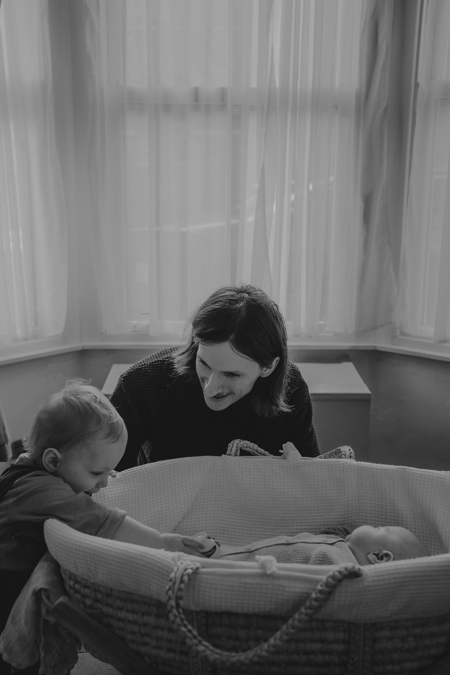 Black and white image of family gathered around bassinet in sunlit room.