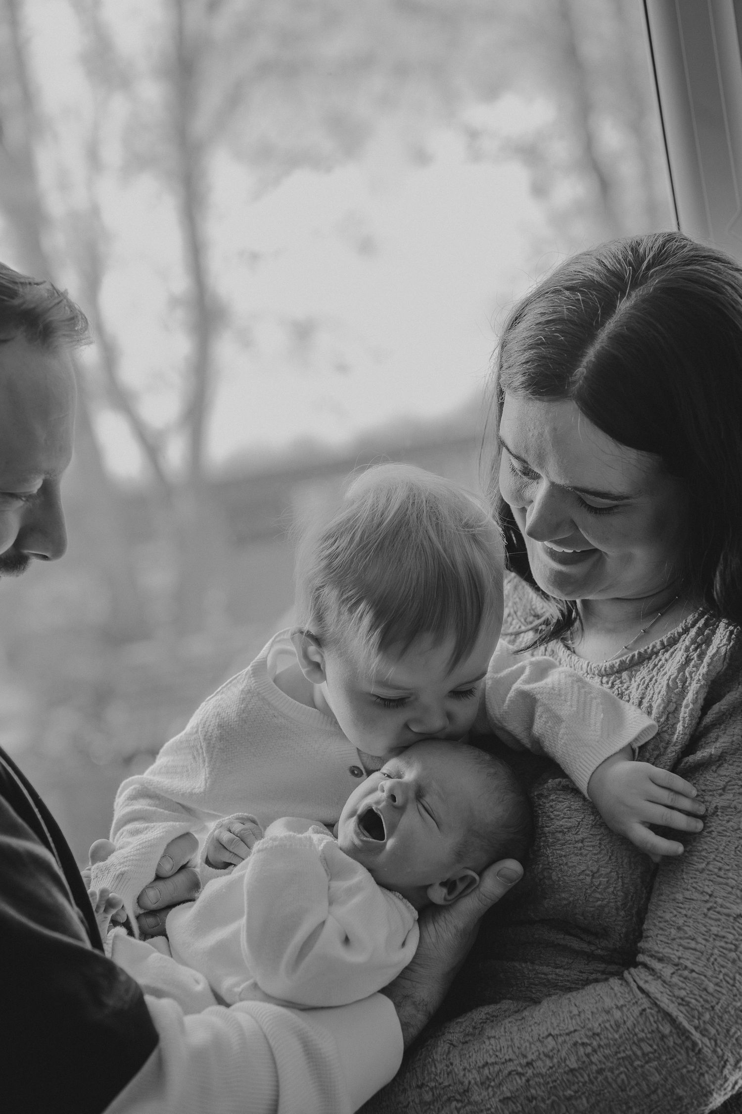 Black and white family portrait of parents with toddler kissing newborn by window.