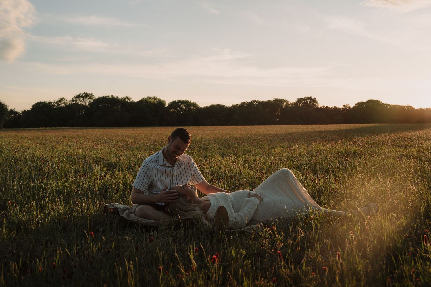 Couple relaxing together in a sunlit green field at golden hour, with trees on the horizon.