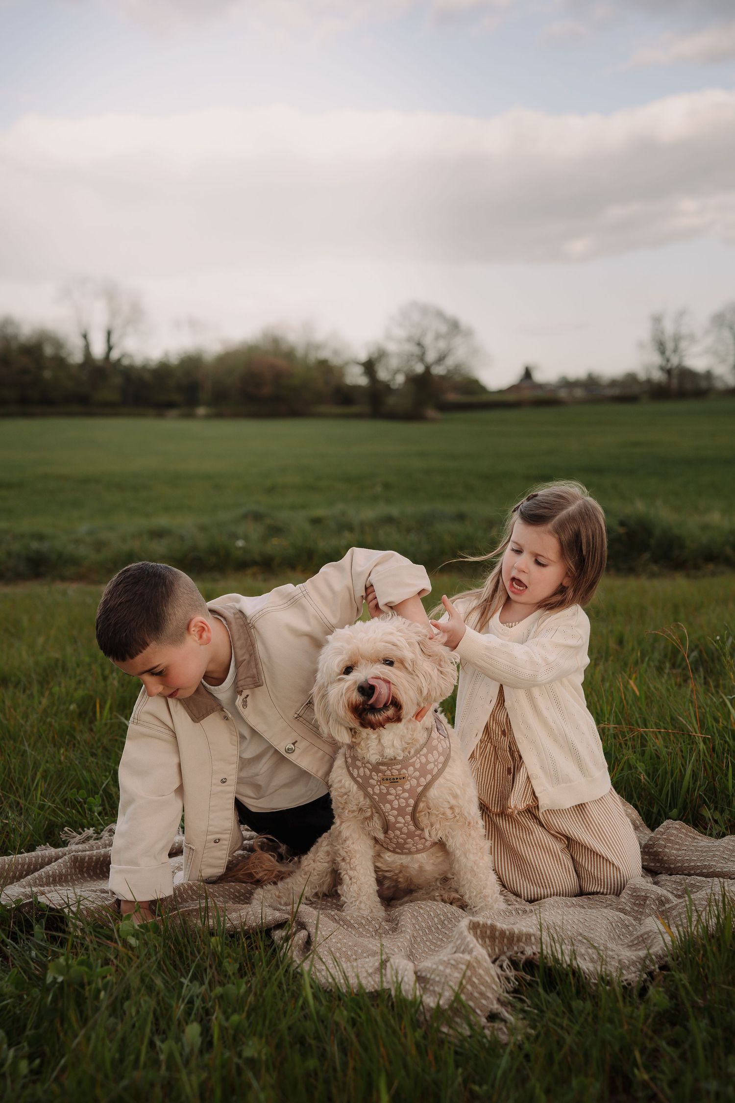 Two children in neutral tones sit on a blanket in a green field with their fluffy cream Goldendoodle dog.
