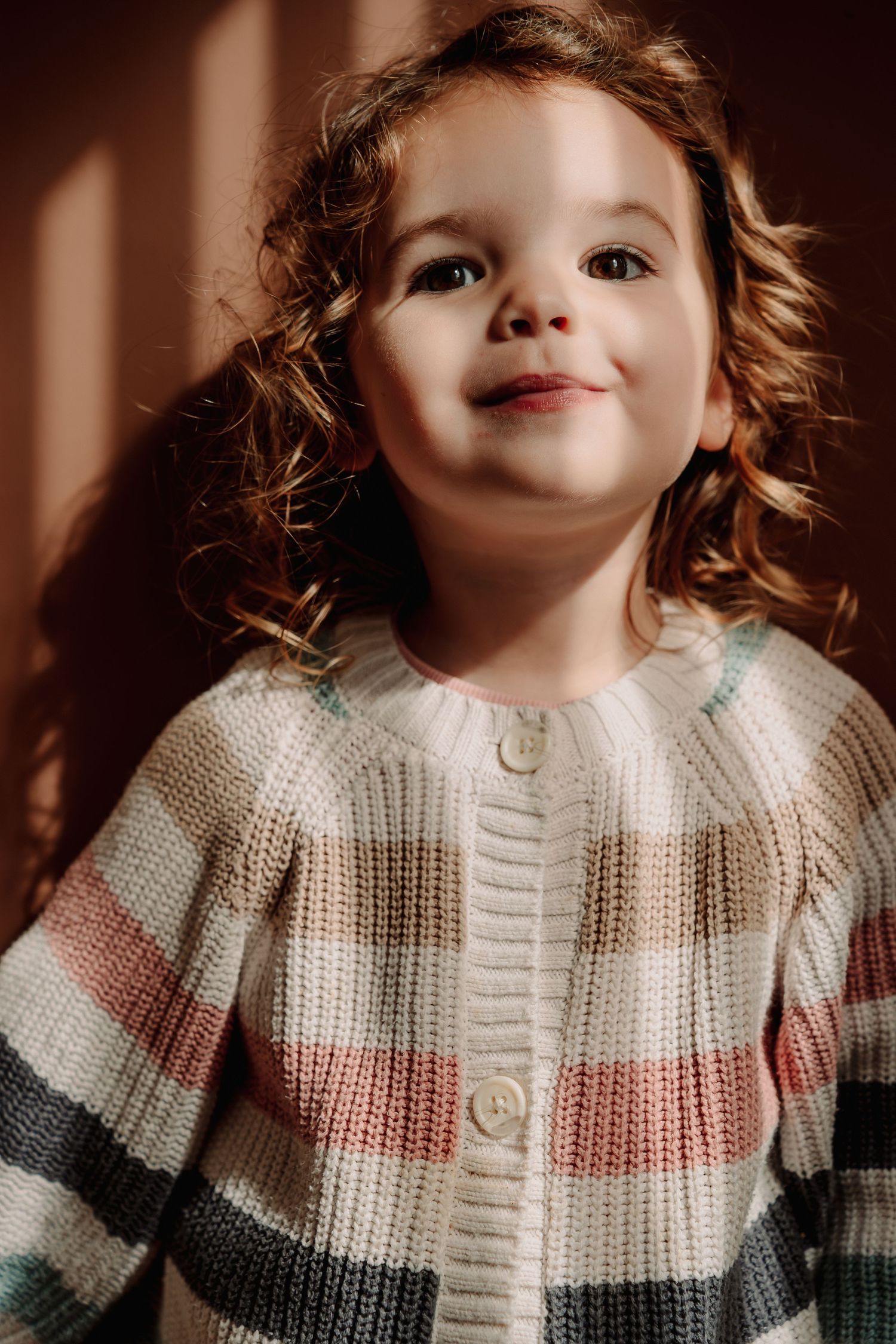 A young child wearing a striped knit cardigan sweater in white, pink and gray stands in warm lighting.