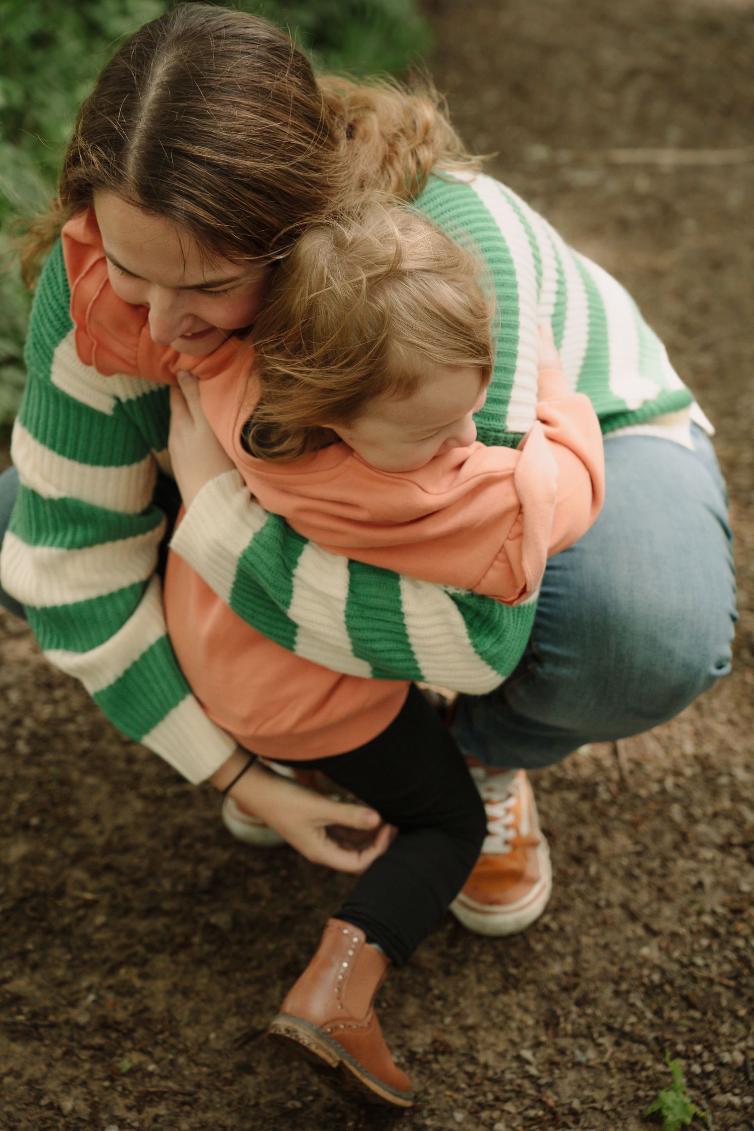 Person wearing green and peach colored sweater crouches down to examine dark soil in a garden or planting area.