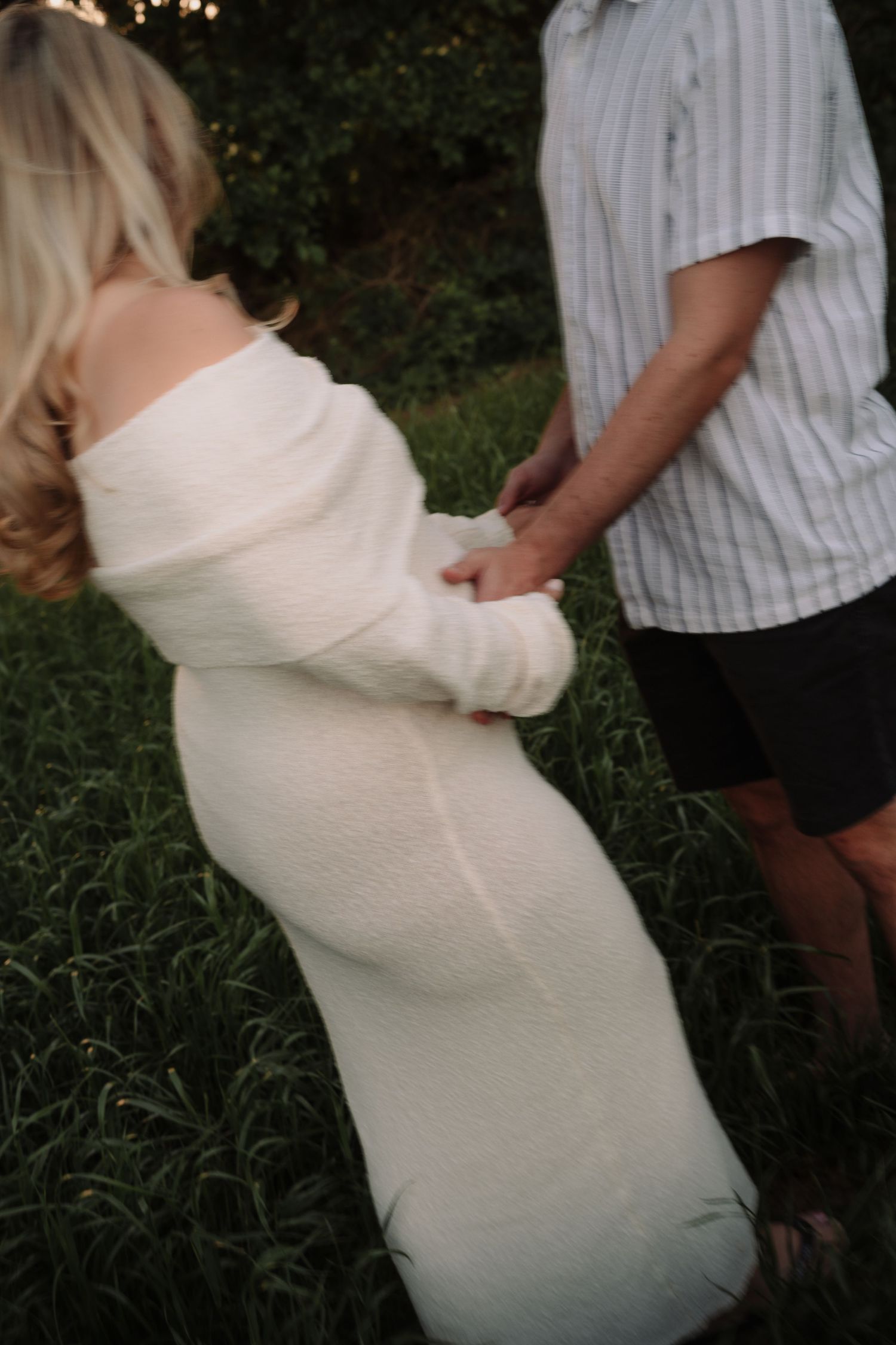 Pregnant woman in white off-shoulder knit dress holds hands with partner in striped shirt outdoors.