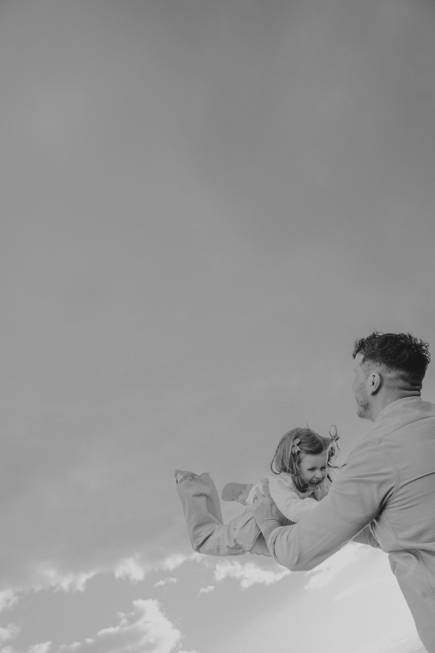 Black and white photo of a father lifting his young daughter against a cloudy sky backdrop.