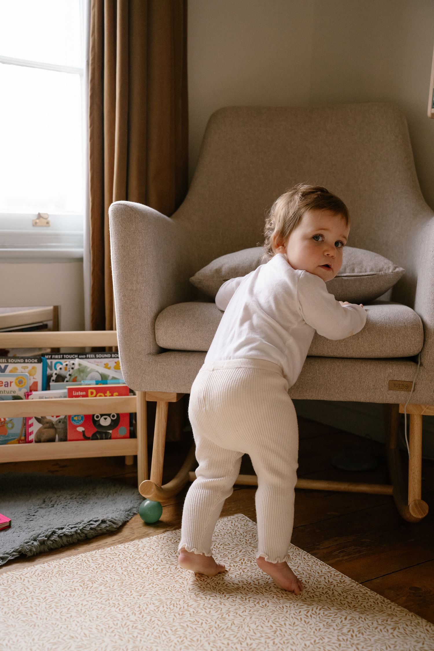 Little girl standing holding onto rocking chair looking back at the camera in an indoor family photo session in Tamworth.