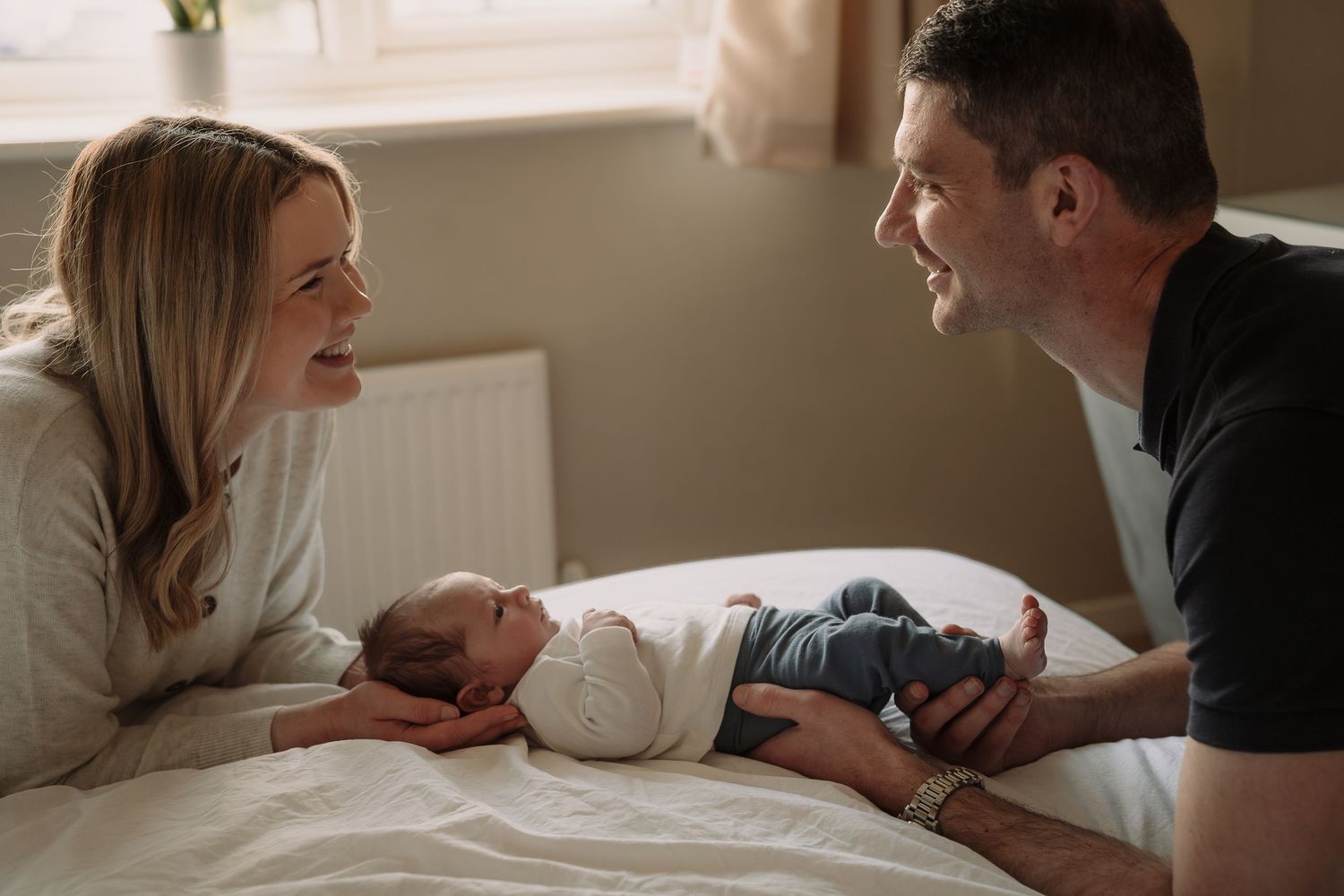Dad and mum leaning on bed with hands on their newborn baby and looking at each other during a Tamworth newborn session .