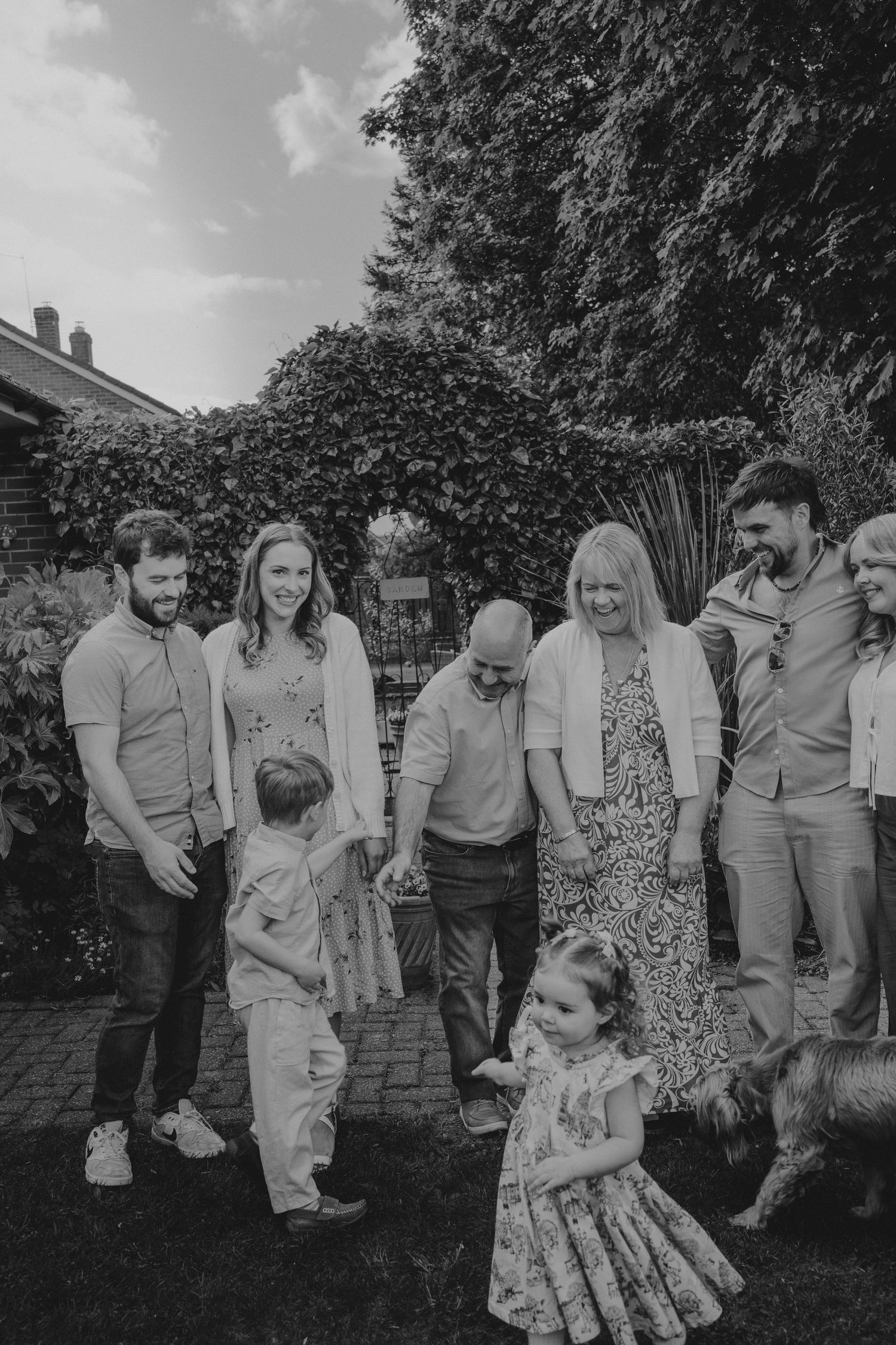 Black and white family photo of multi-generational group gathered outdoors in garden with two young children and a dog.