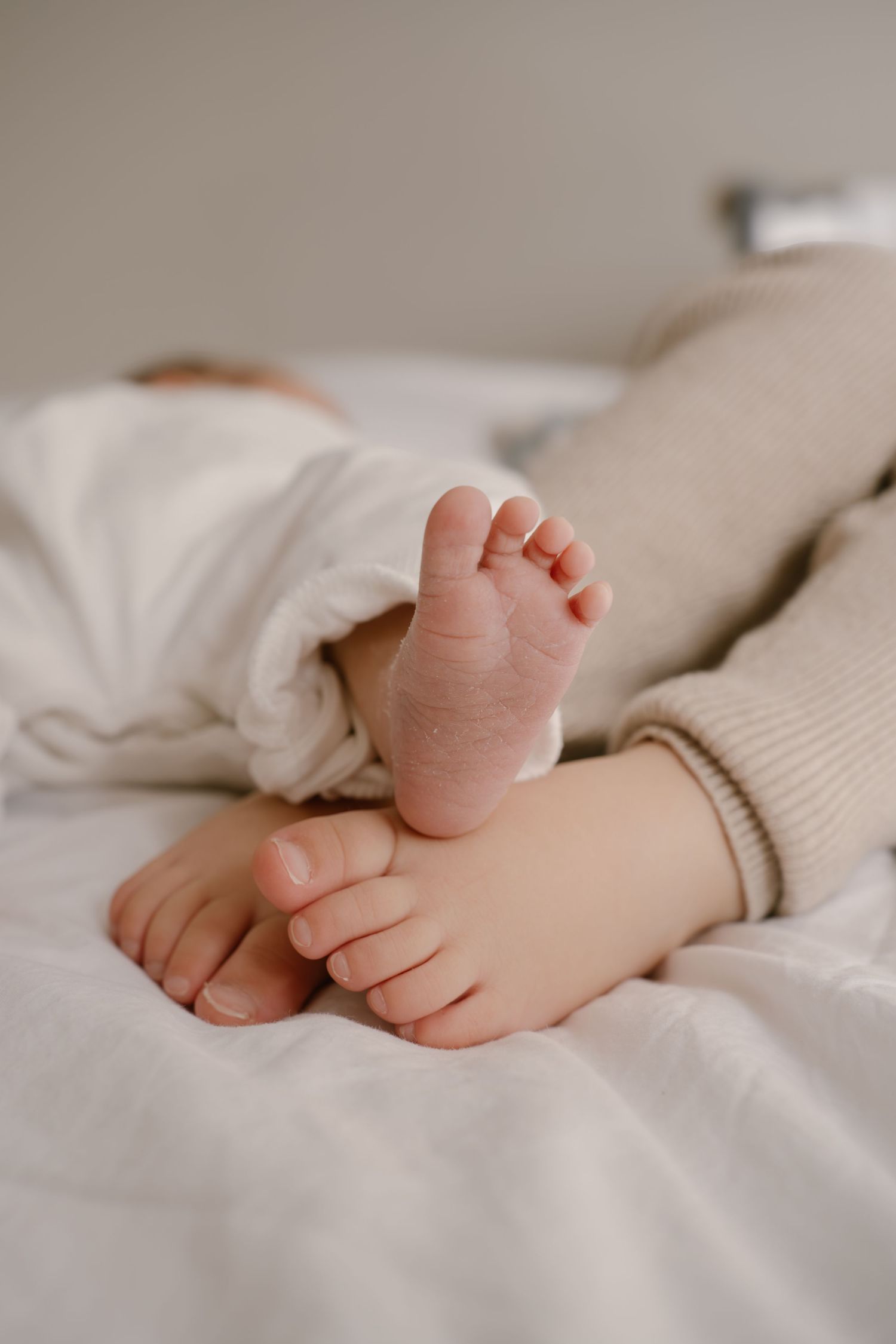 Tiny baby feet peek out from soft white bedding showing delicate toes against a neutral background.