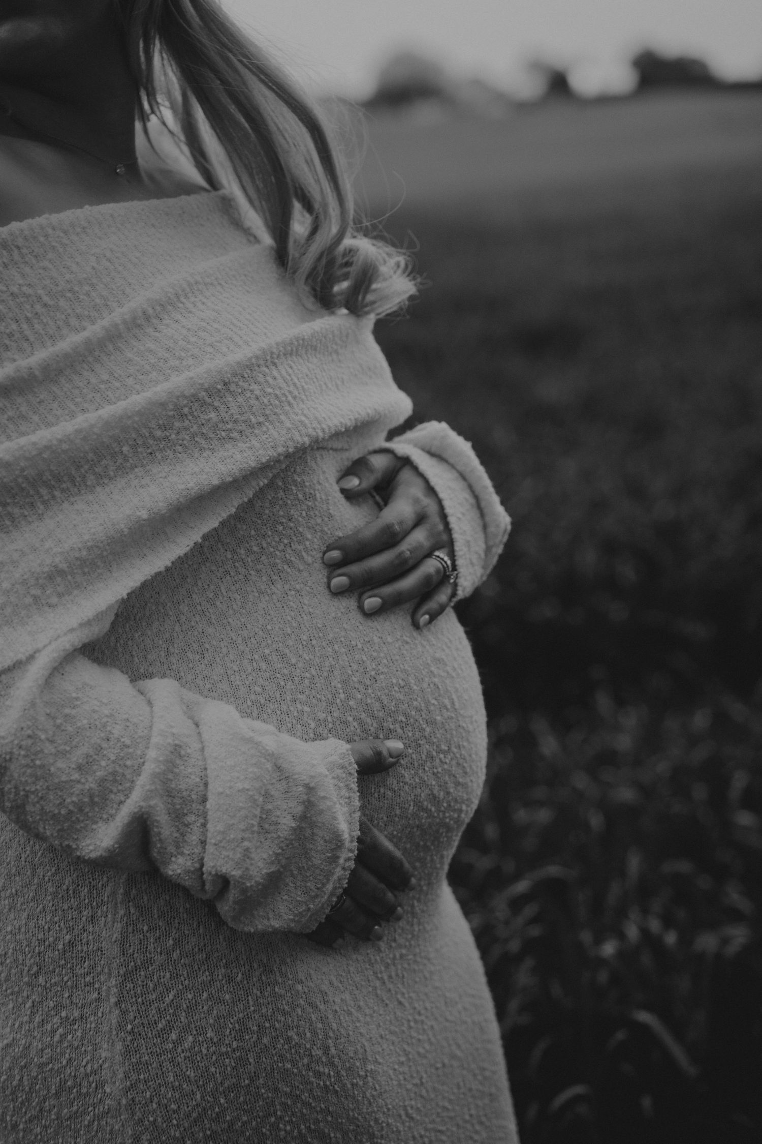 Black and white photo of a pregnant woman in a cozy sweater cradling her baby bump outdoors in a field.