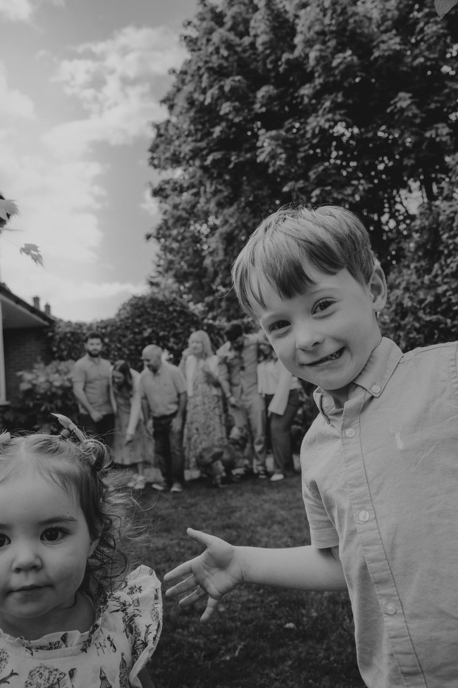 Black and white photo of two young children smiling at a garden party with adults gathered in the background.