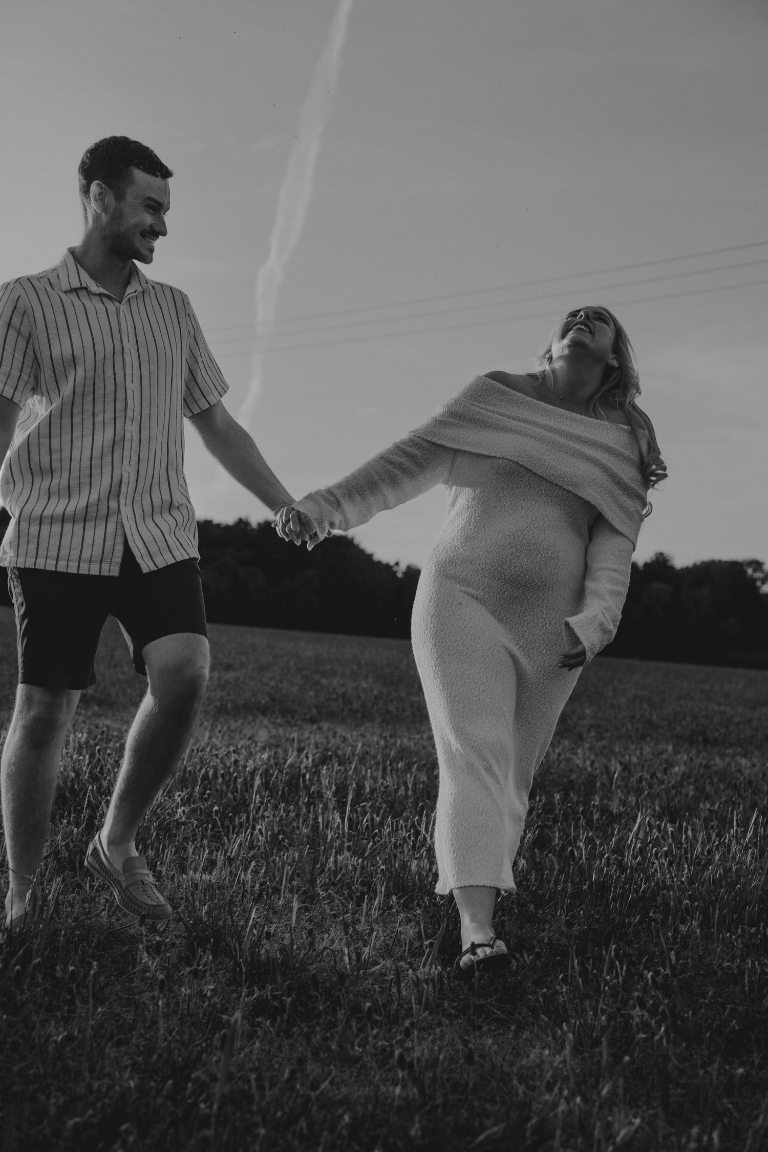 Black and white photo of a pregnant couple holding hands and laughing in a grassy field at dusk.