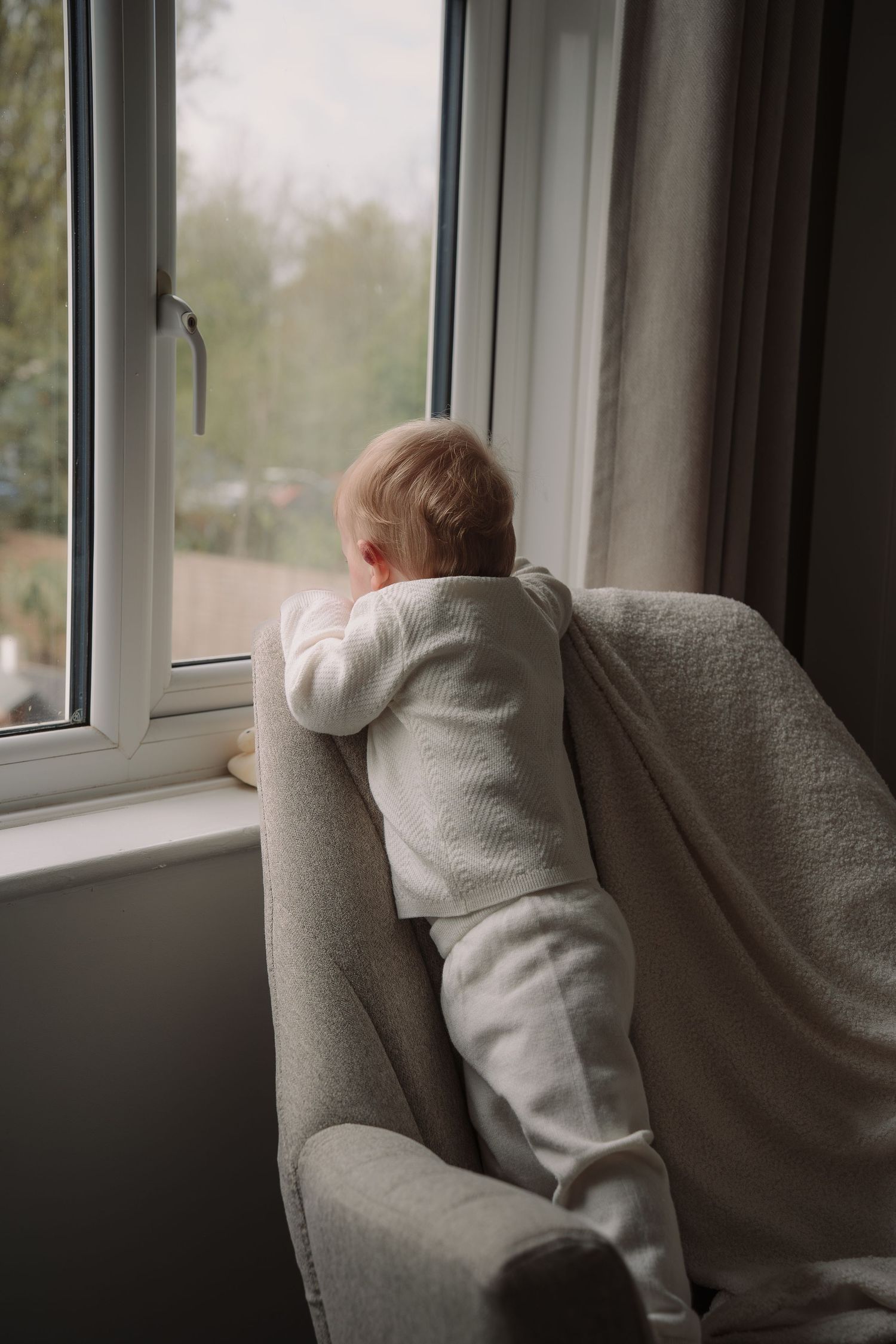 Baby in white onesie standing on armchair looking out window.