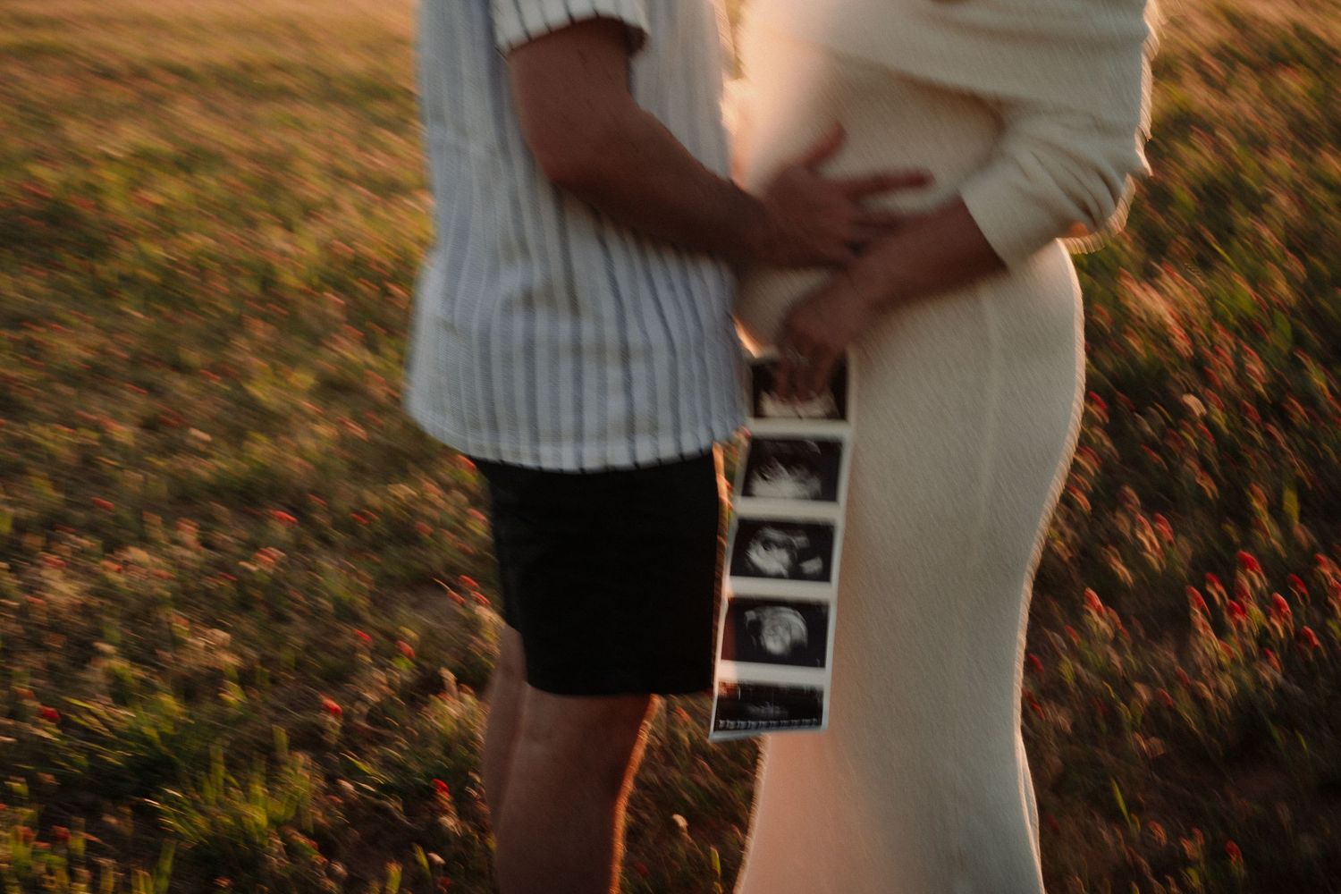 Couple holding ultrasound photos outdoors at golden hour, pregnant woman in white dress embracing partner in striped shirt.