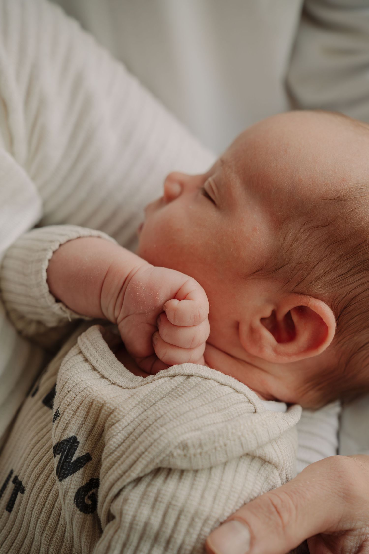 A sleeping newborn baby wearing a cream-colored knit sweater rests peacefully in a soft, natural light setting.