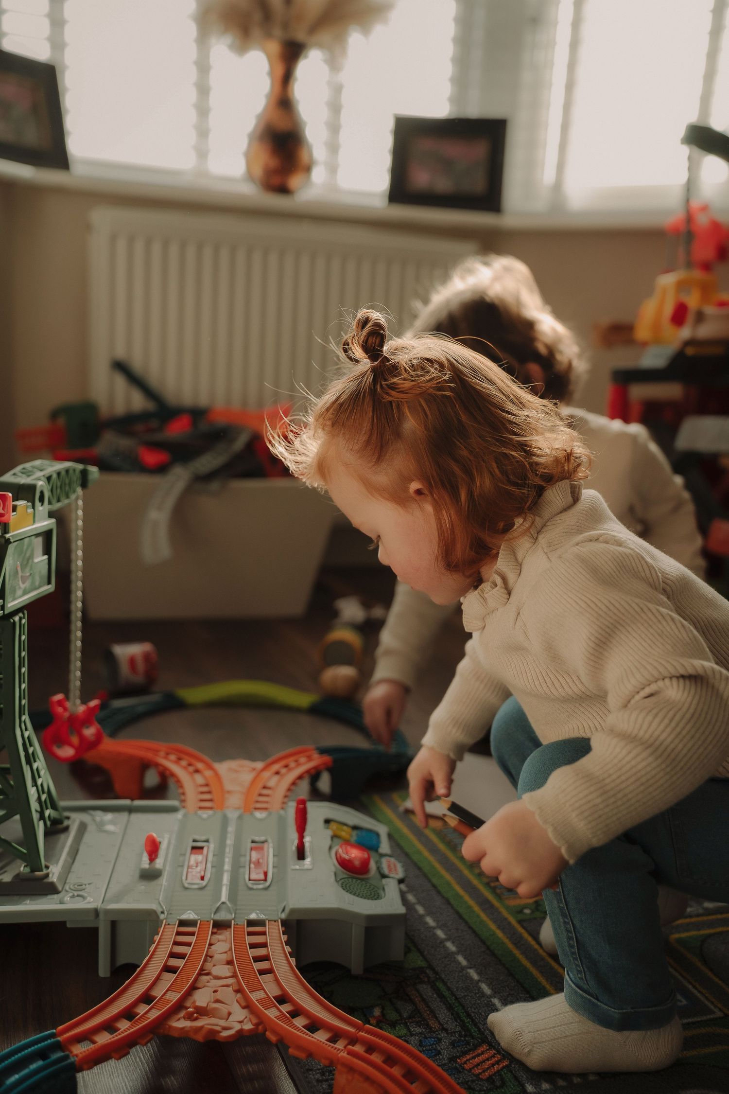 A child in a cream sweater plays with a toy train track set on a wooden floor in a living room setting.