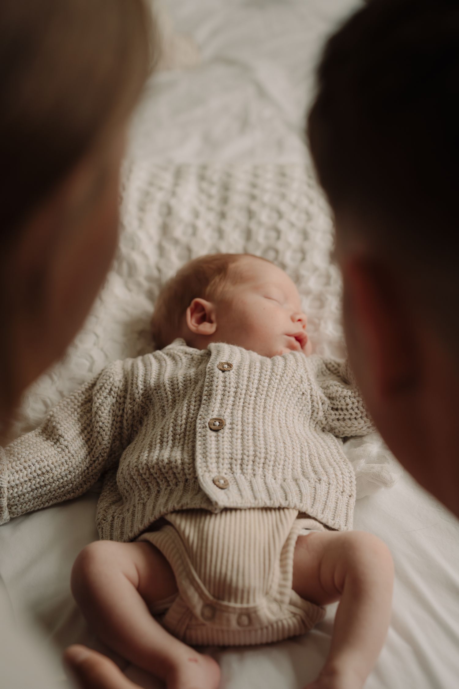 Baby wearing knit sweater lying on white bedding in soft lighting.
