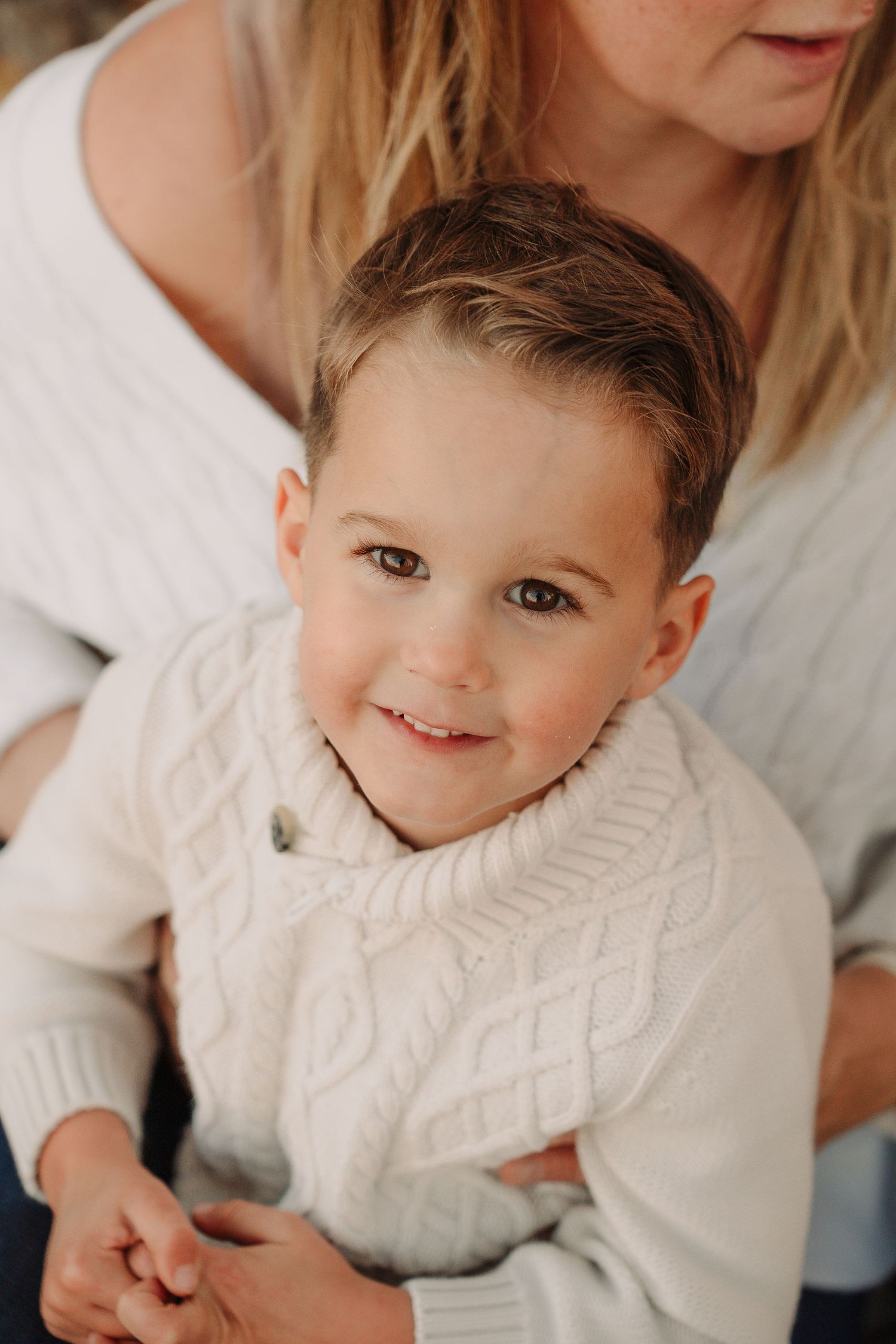 A young child in a white cable knit sweater smiles warmly while sitting with mum During a family photoshoot by a Tamwoth photographer.
