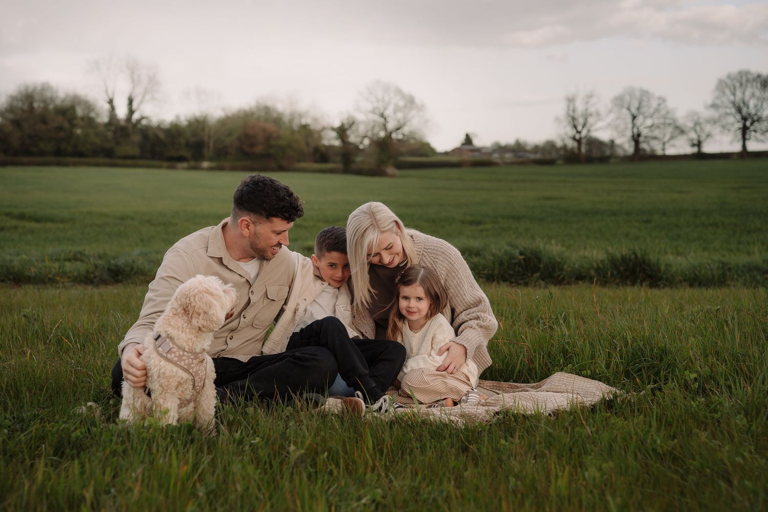 Family with two children and a dog sitting on a blanket in a lush green field during a calm evening.
