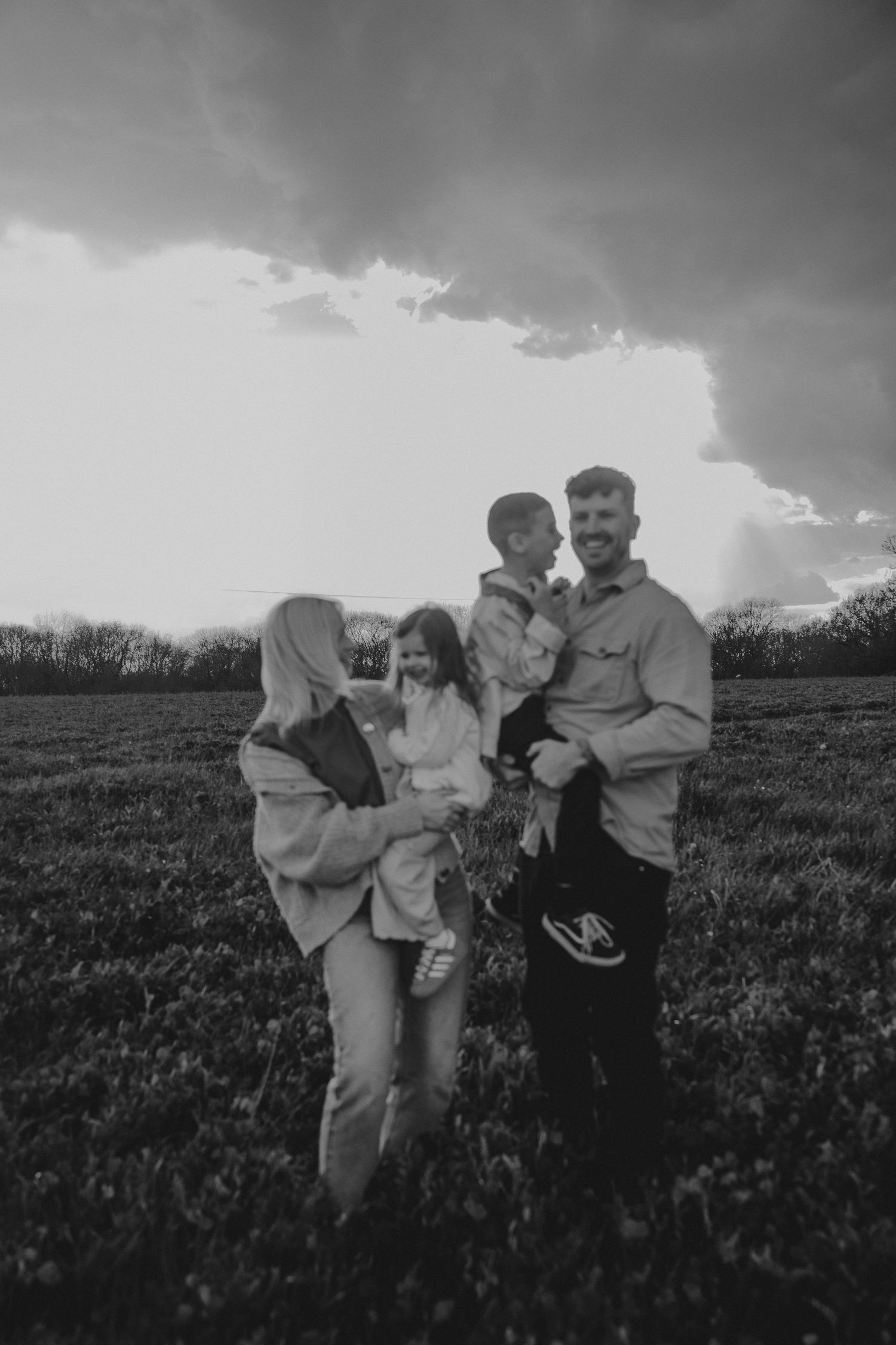 Black and white photo of a happy family of four posing in a field under dramatic storm clouds at dusk.
