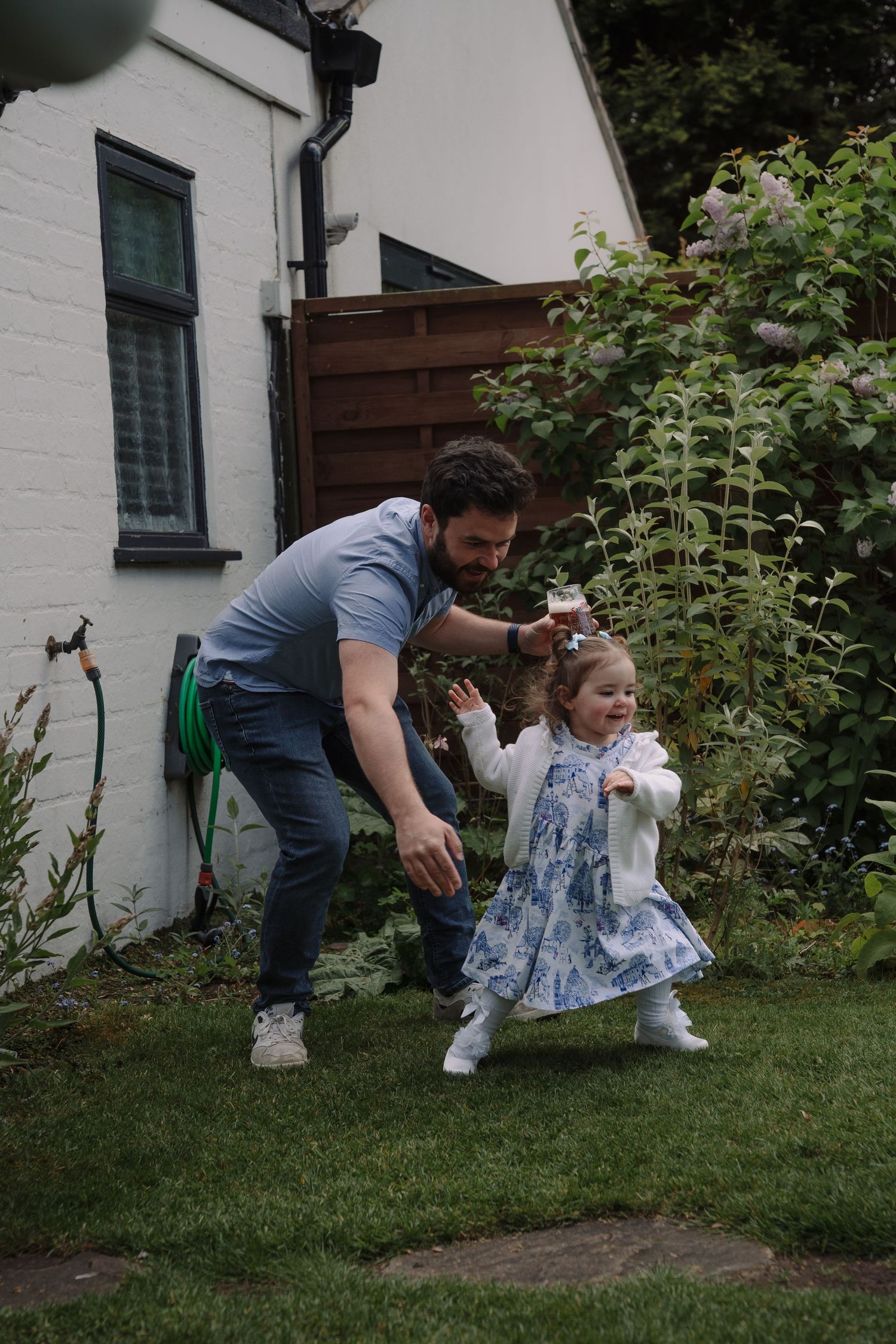 Father helps toddler girl walk in garden, child wearing blue floral dress and white cardigan, playing outdoors near house.