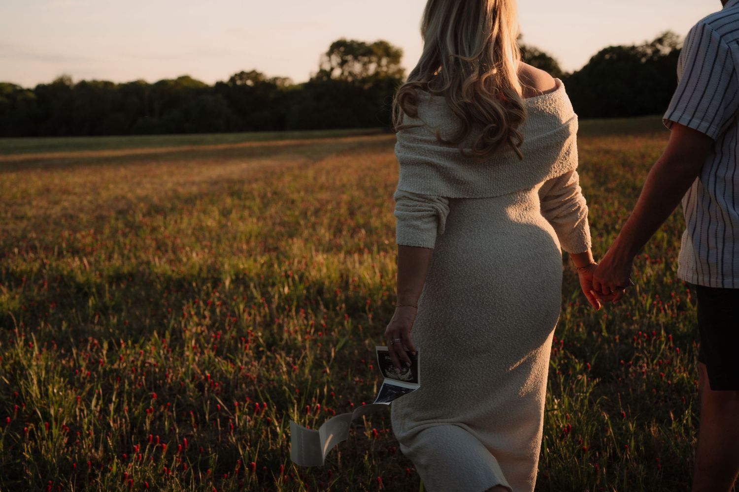 Pregnant woman holding ultrasound scan hands with partner in golden sunset field, maternity photography outdoors.