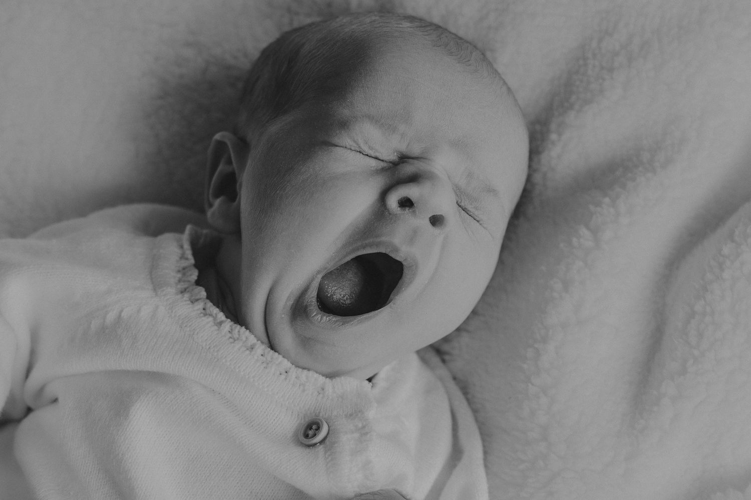 Black and white photo of a newborn baby yawning while lying on a soft blanket.
