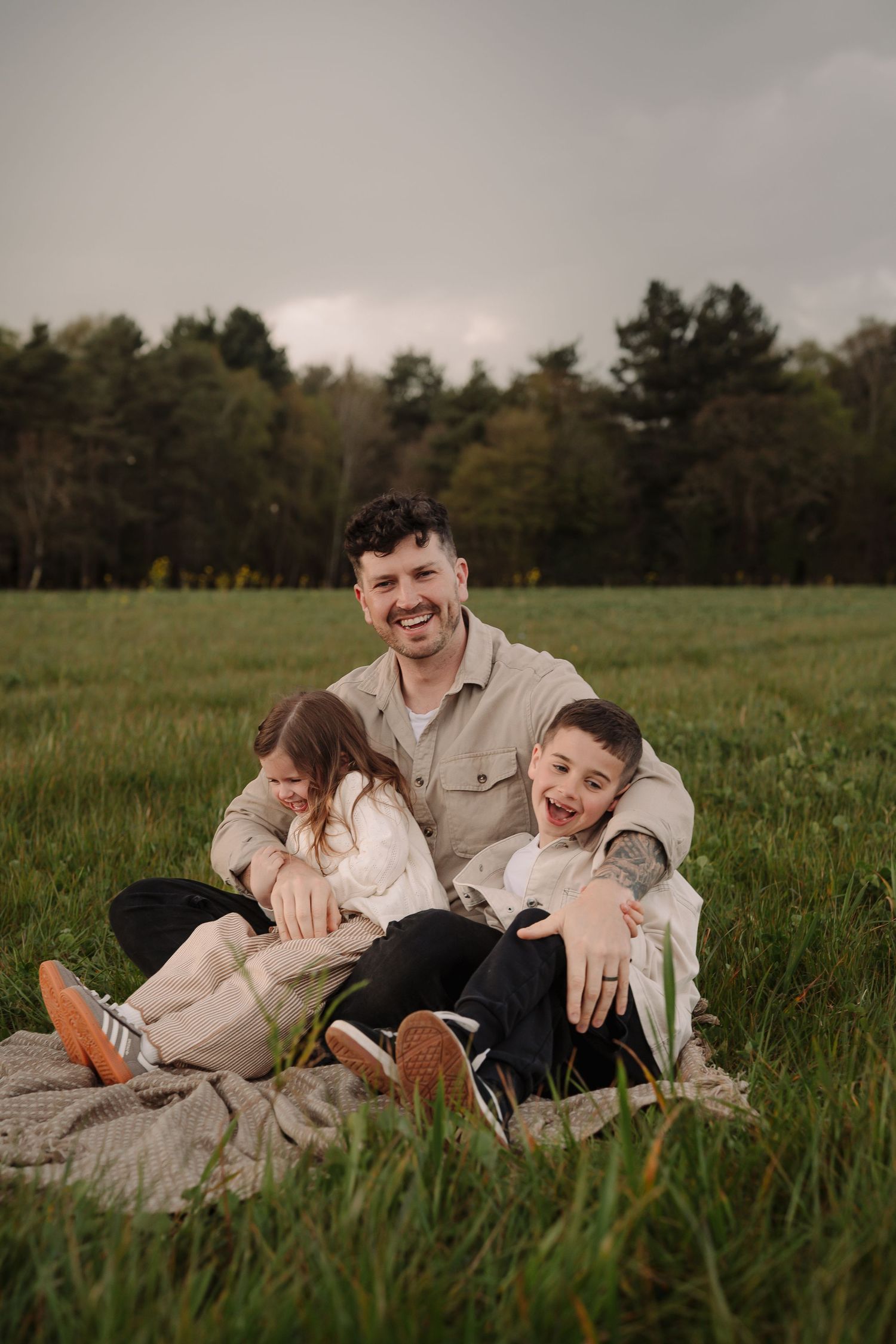 Smiling father sitting on a blanket in a green field with his young daughter and son leaning against him.