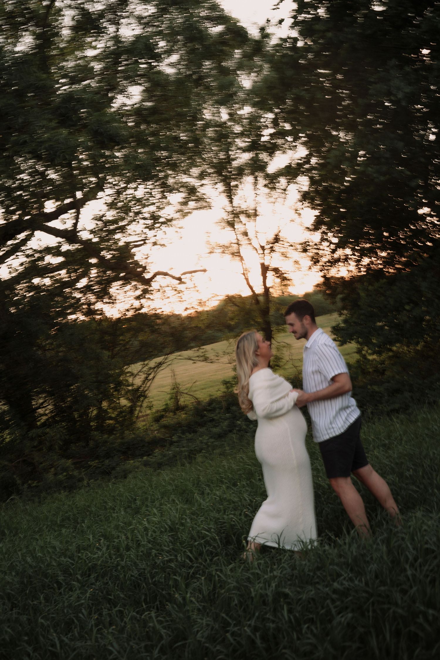 Couple embracing at sunset in a lush green field, woman in white dress, golden hour glow behind trees.