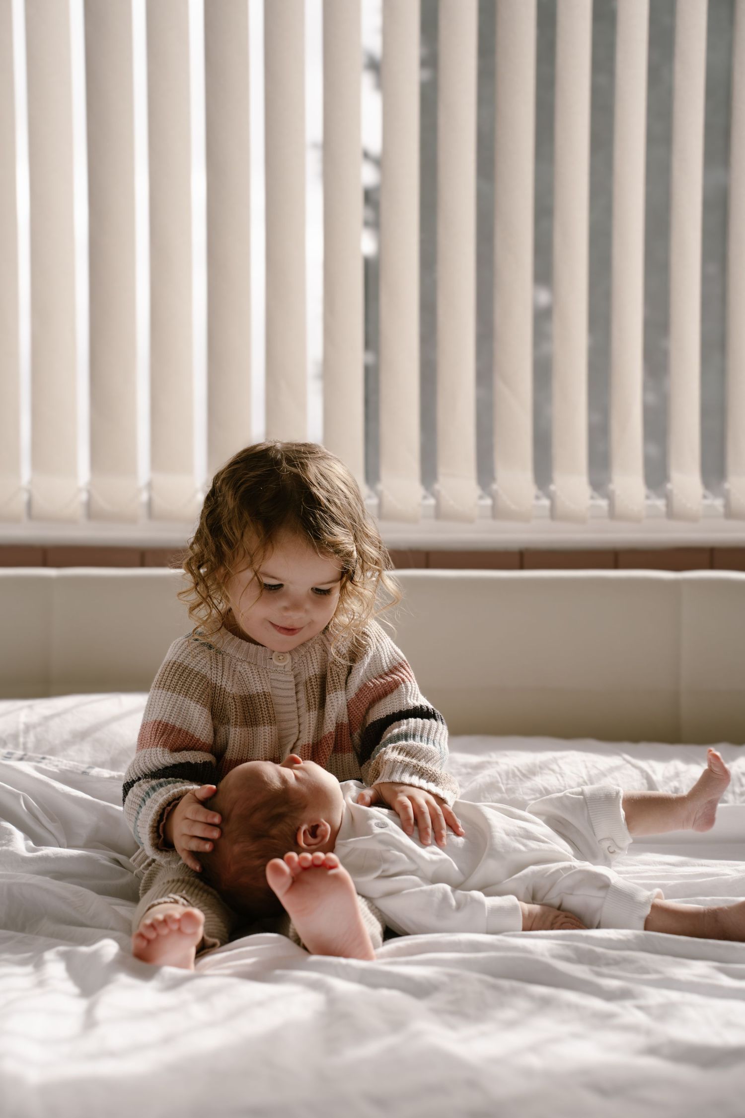 A small child in a knit sweater sits on white bedding near a window with vertical blinds.
