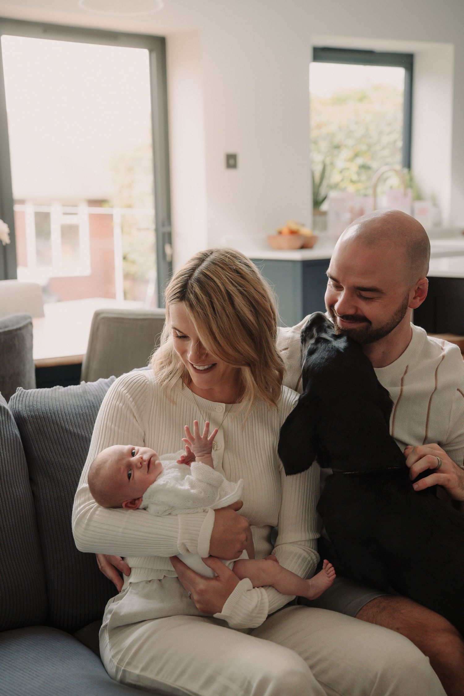 A family shares tender moments together on a gray couch in a series of candid lifestyle photographs.