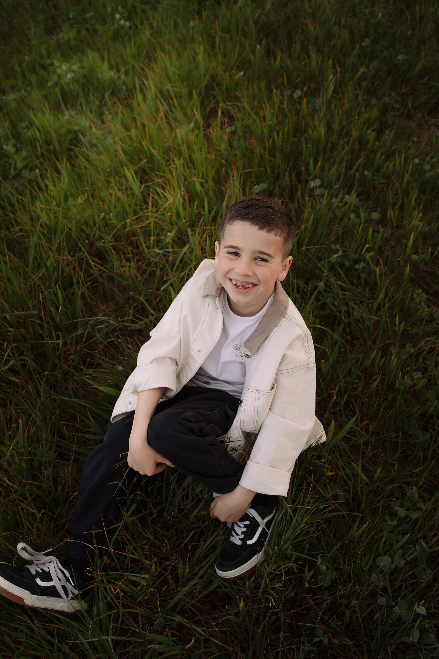 Smiling boy sitting in green grass wearing a white jacket, black pants, and Vans sneakers.