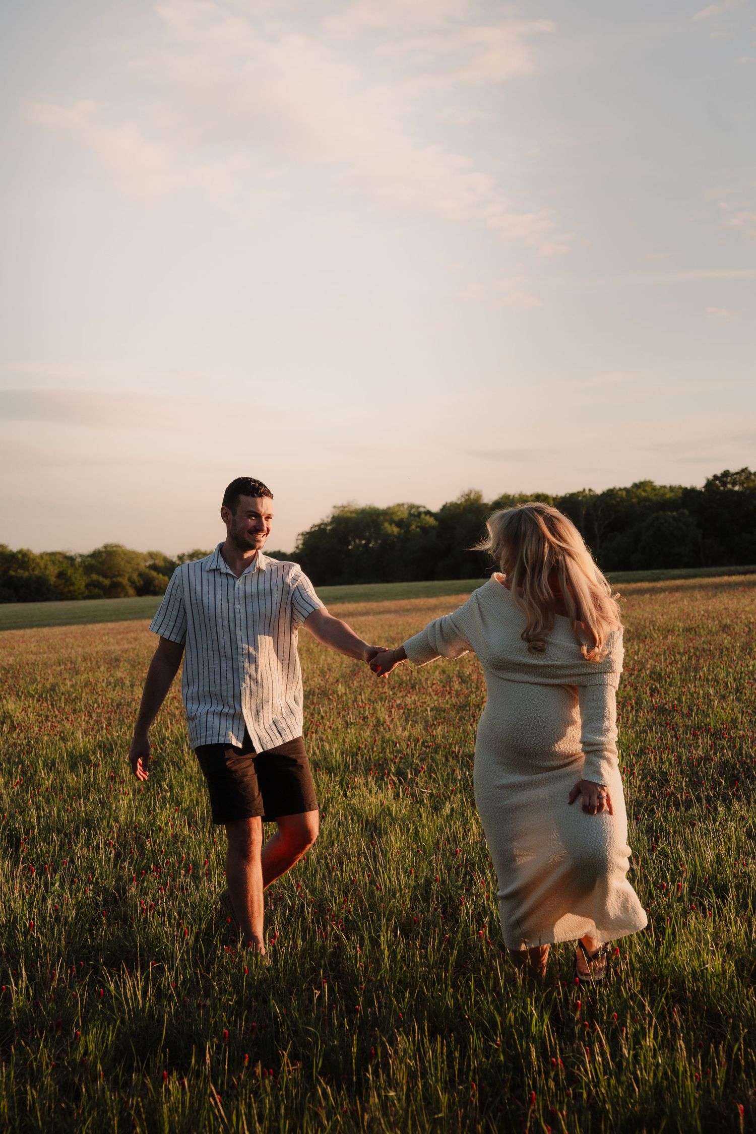 A couple holds hands while walking through a lush green field at golden hour, the woman in a flowing white dress.