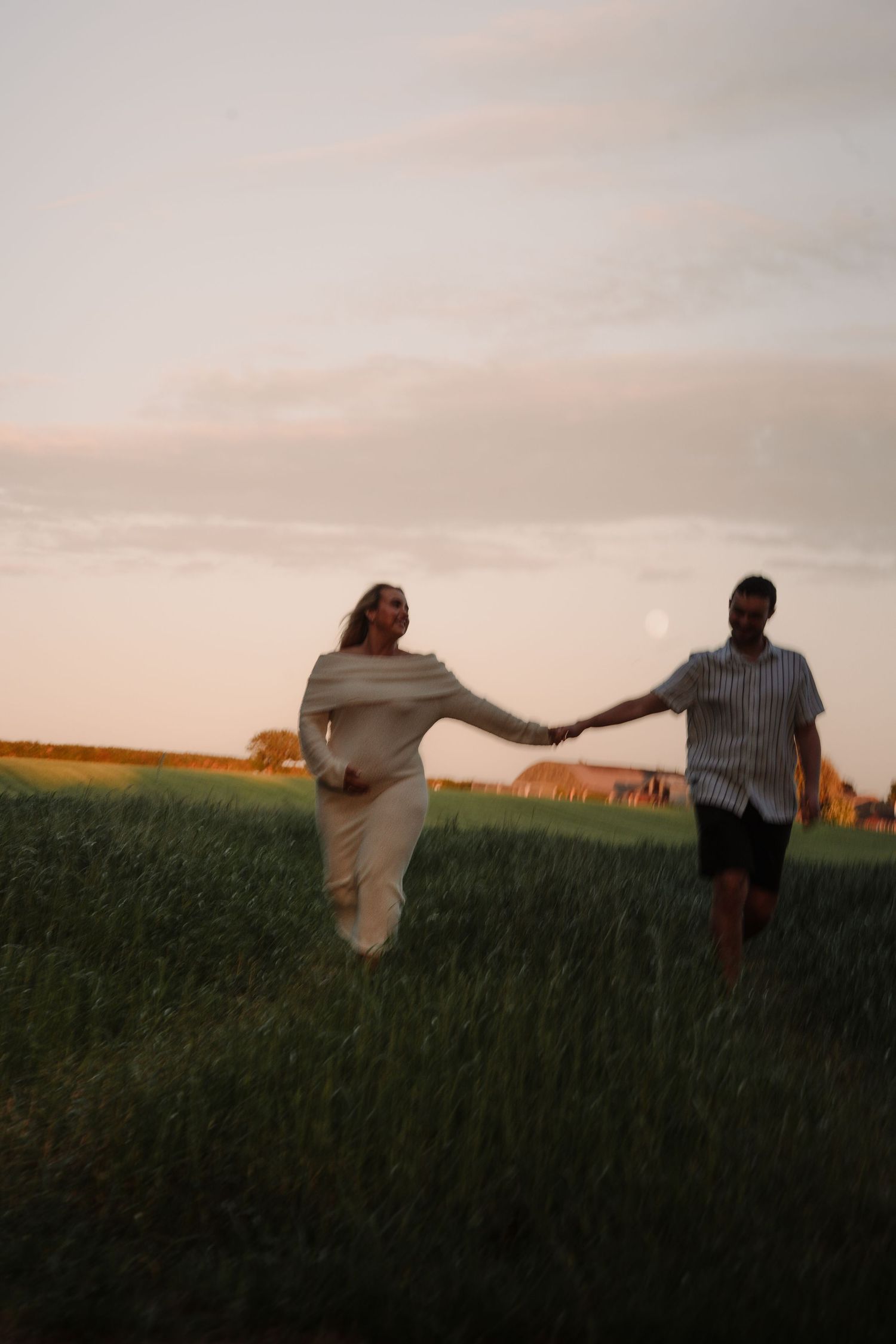 Couple holding hands walking through green field at golden sunset, woman in white dress, man in striped shirt.