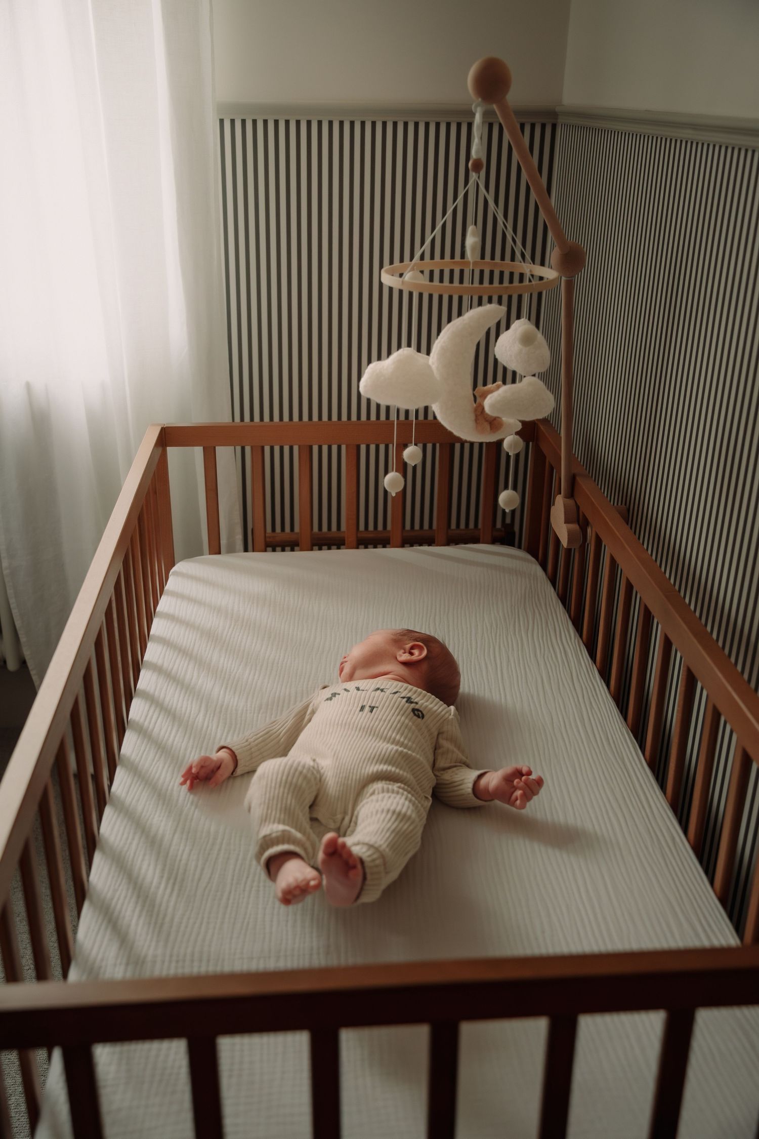 Baby wearing white onesie lies in wooden crib with mobile hanging above featuring plush animals and toys.