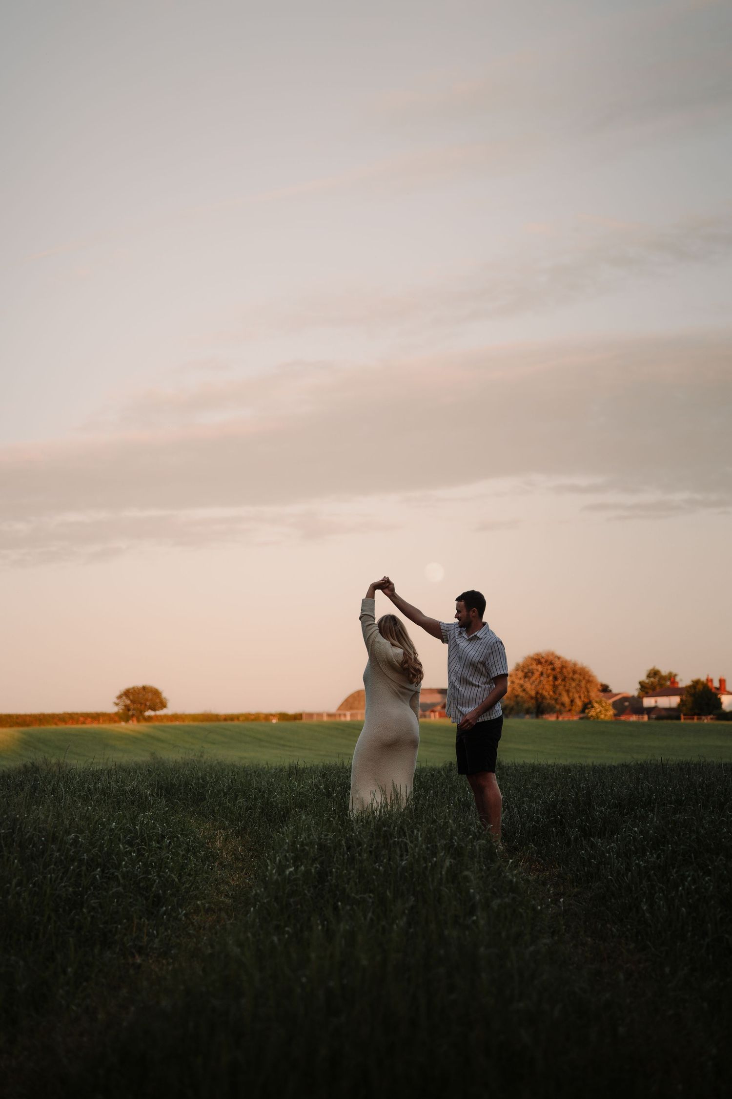 Couple dancing in a green field at golden hour sunset, woman in white dress, romantic outdoor photography.