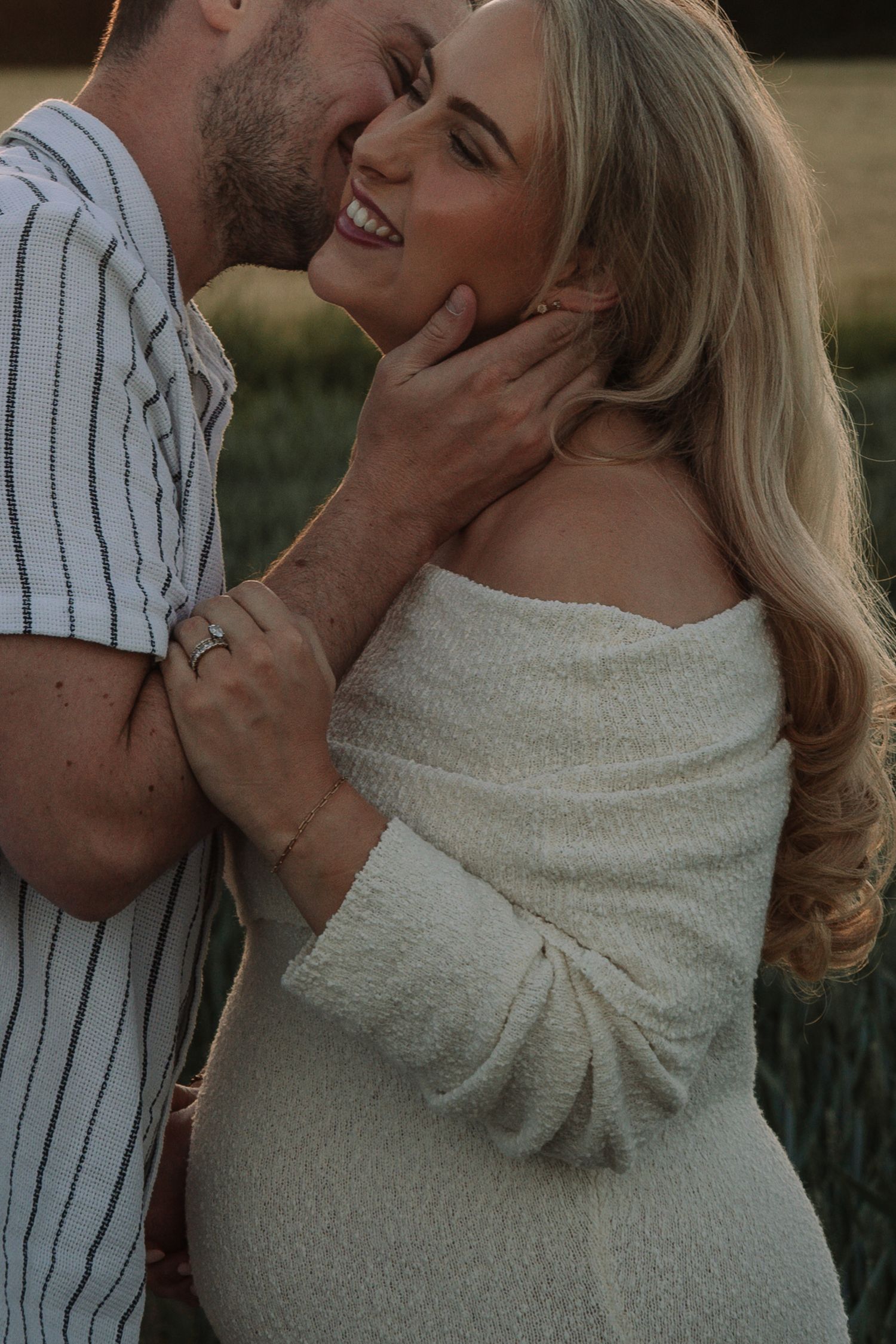 Smiling pregnant woman in white off-shoulder dress embraced by man in striped shirt during golden hour outdoor maternity shoot.
