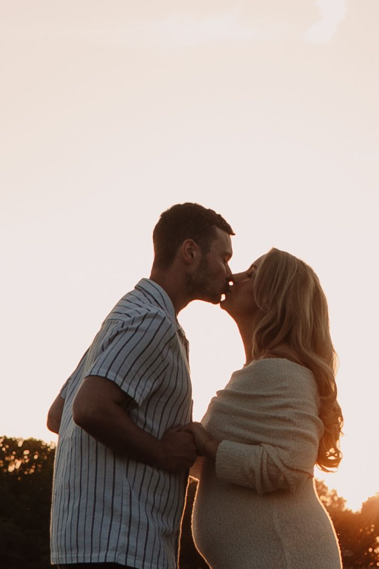 A couple shares a romantic kiss at golden hour sunset, the man in a striped shirt embracing a blonde woman in a cream sweater.
