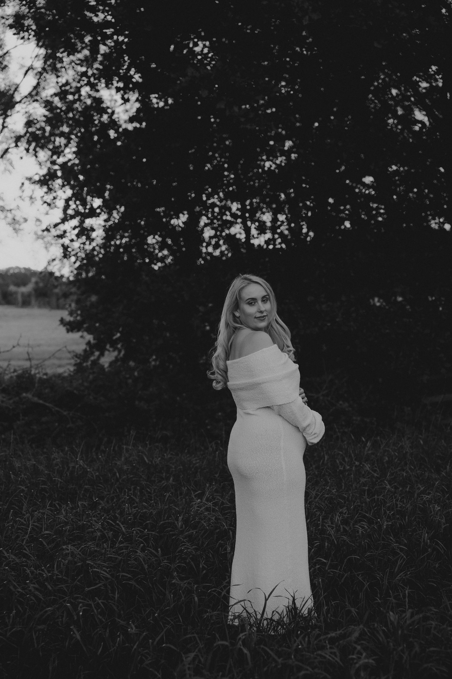 Black and white photo of a blonde woman in an elegant off-shoulder white gown standing in a lush outdoor field.