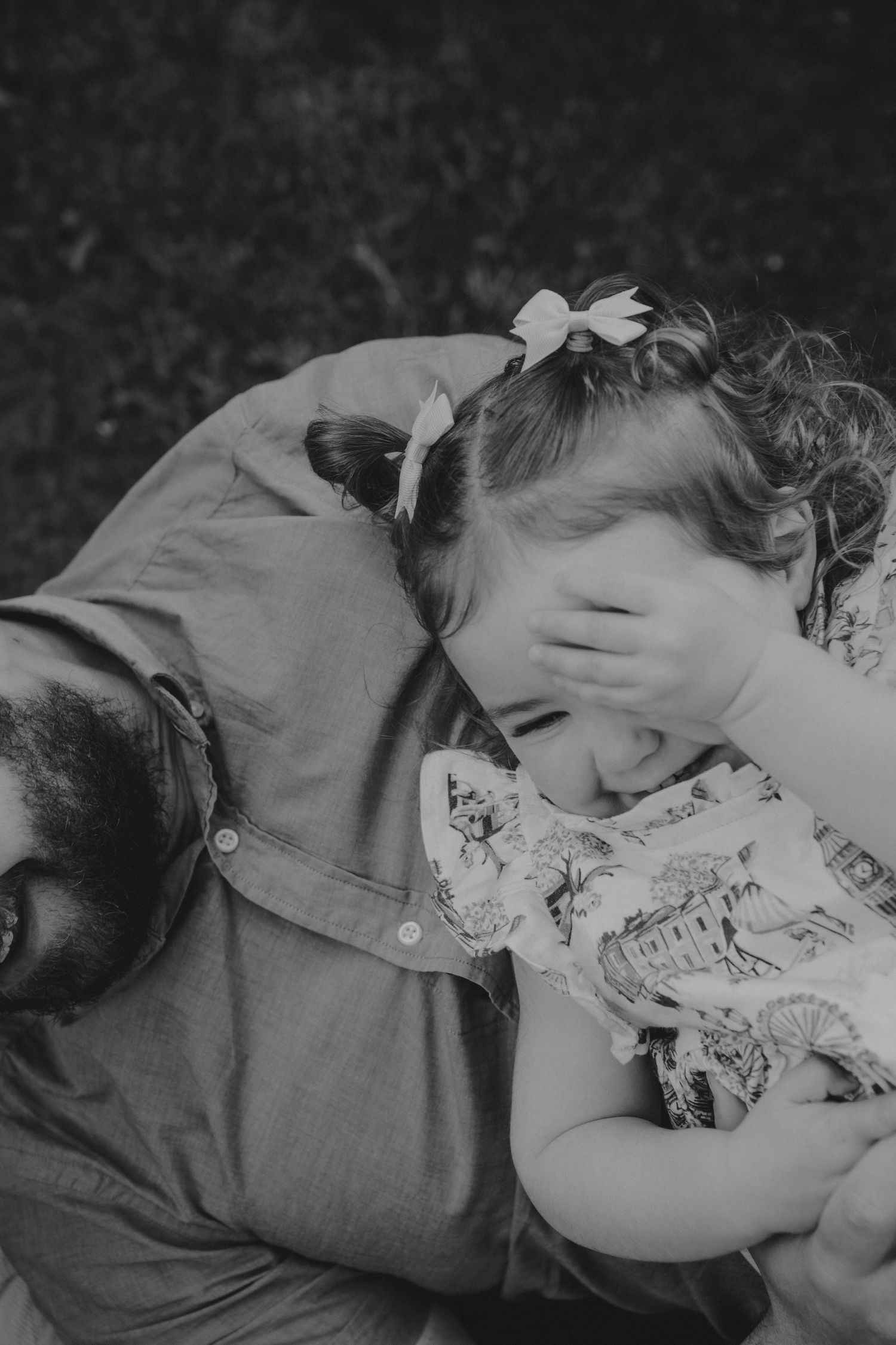 Black and white photo of a laughing toddler girl with bow pigtails cuddling with her bearded father outdoors.