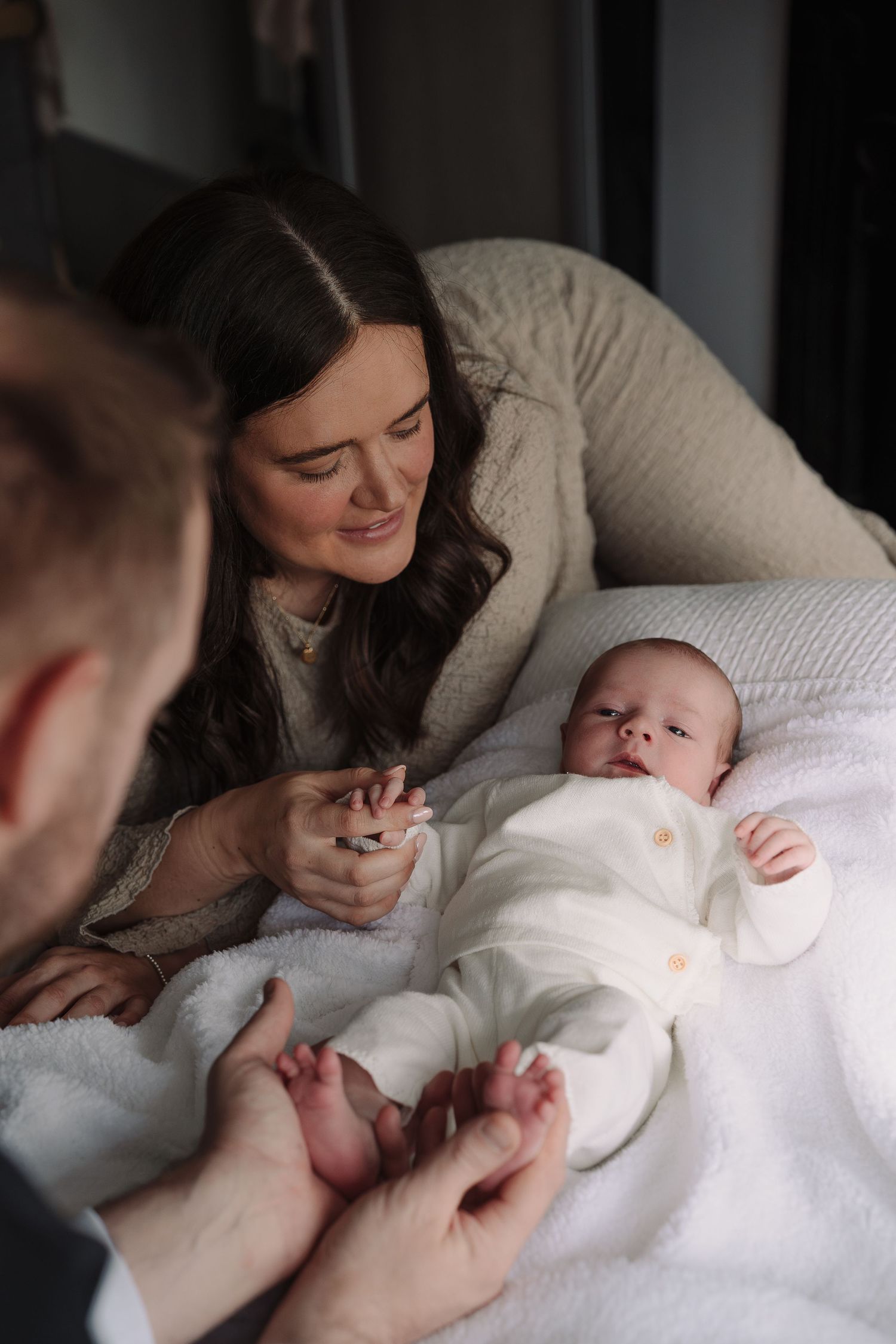 A mother smiles lovingly at her newborn baby in white, while a father gently holds the infant's feet on a soft blanket.