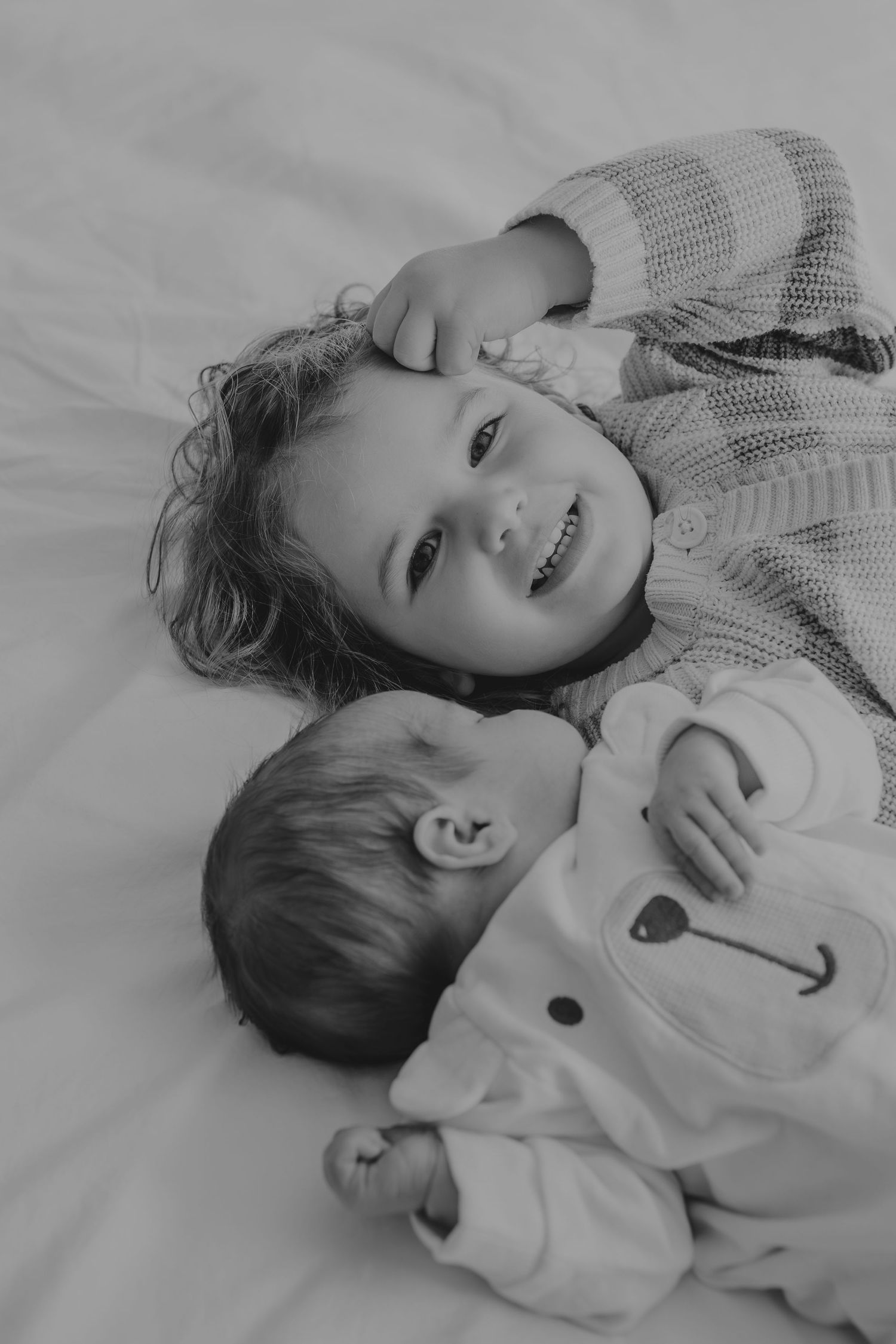 Black and white portrait of two children lying together on white bedding, sharing a tender moment.