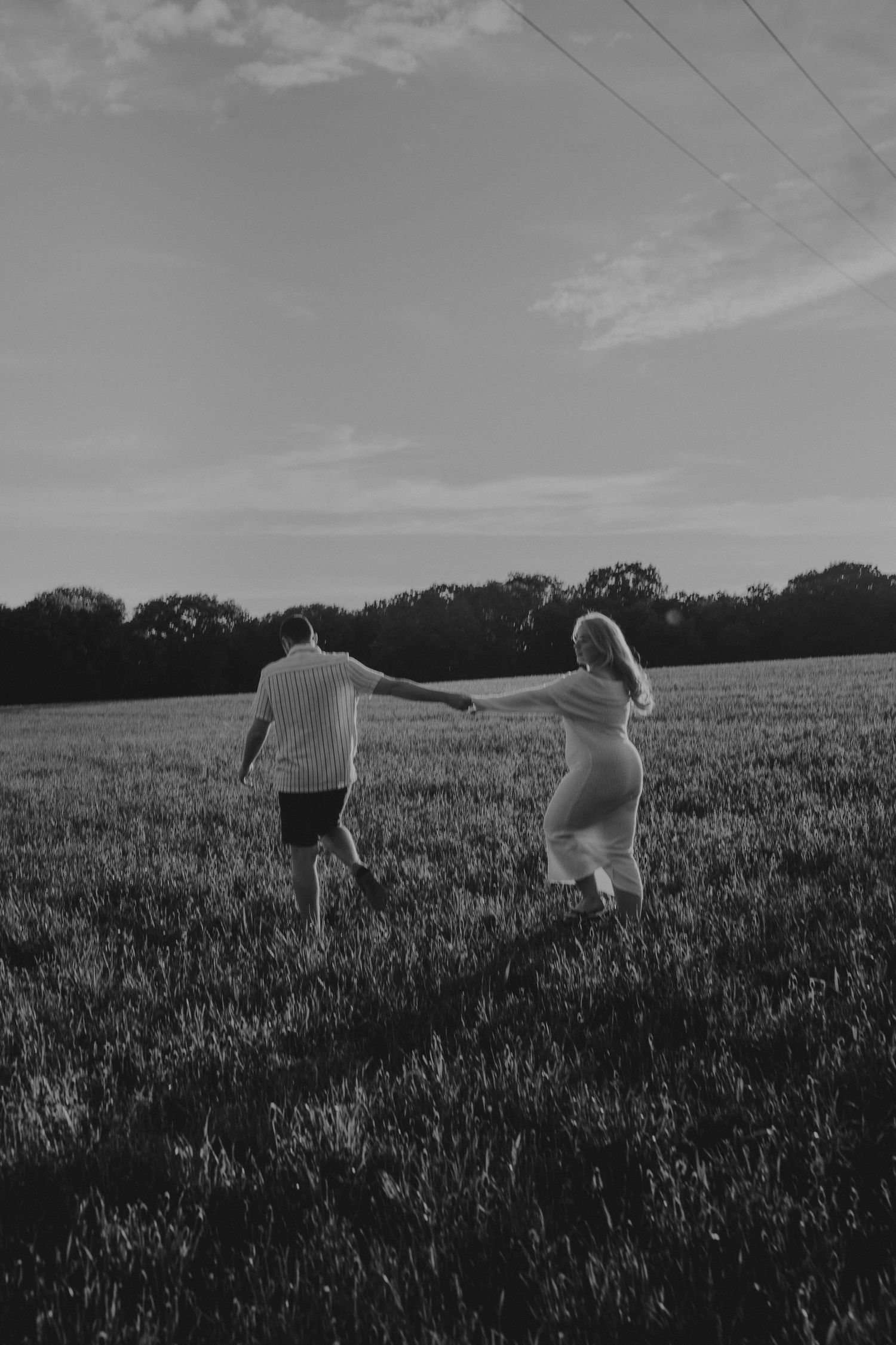 Black and white photo of a pregnant couple holding hands while walking through a lush green field at sunset.