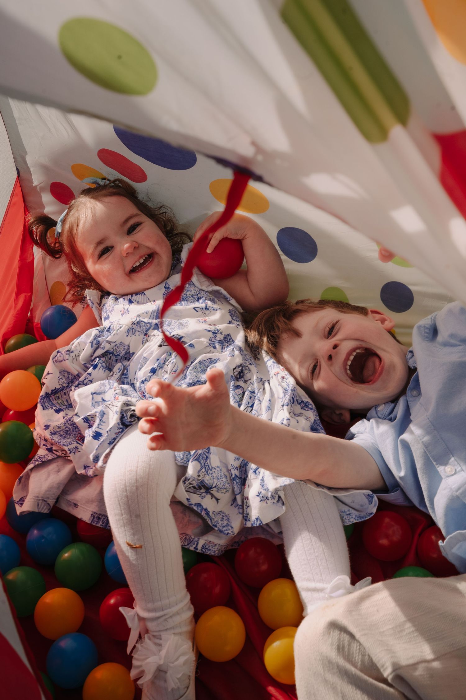 Two laughing children playing in a colorful ball pit under a polka dot play tent, full of joy.
