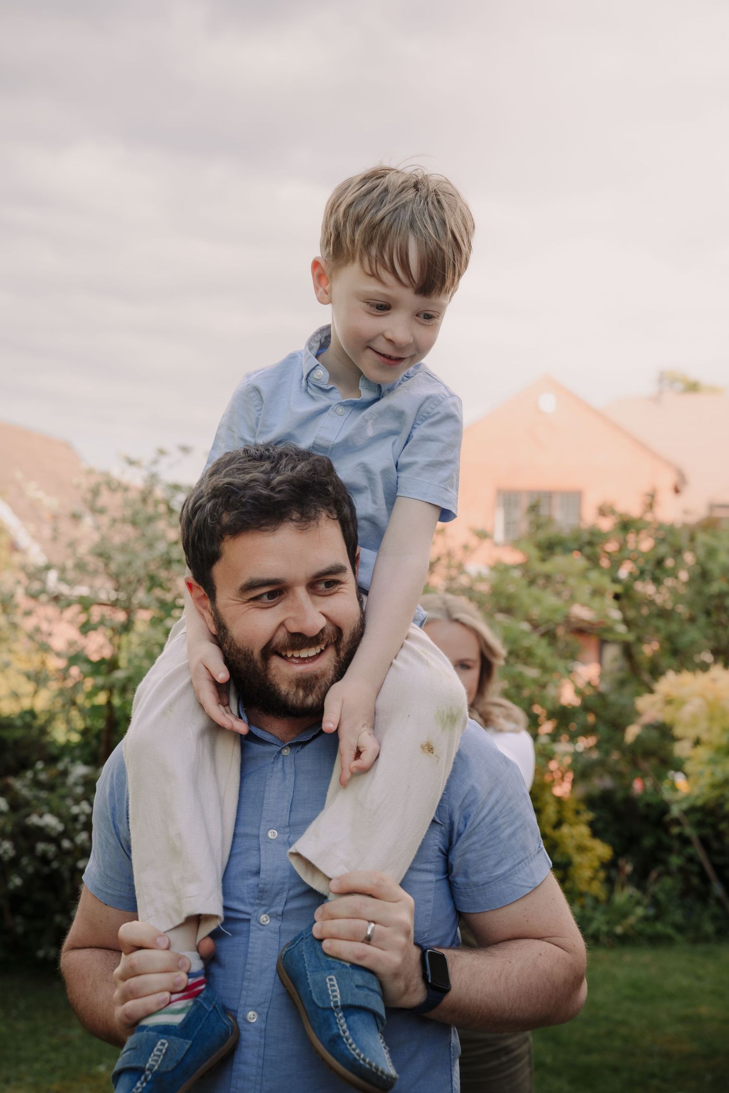 A smiling man carries a young blonde boy on his shoulders in a garden, with a woman visible behind them.