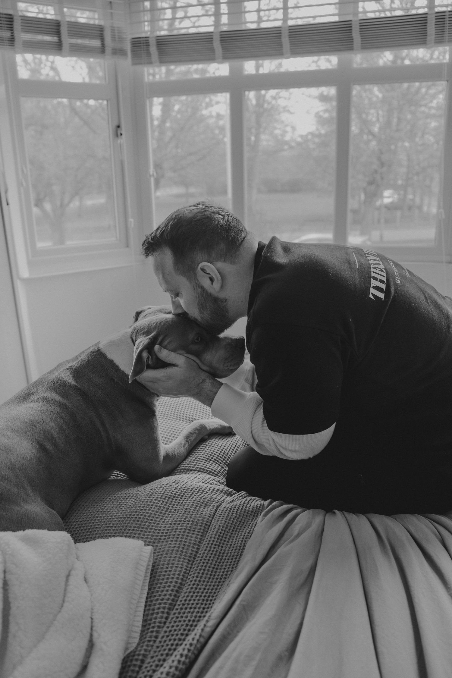 Black and white photo of a bearded man tenderly kissing his dog by a bright window.