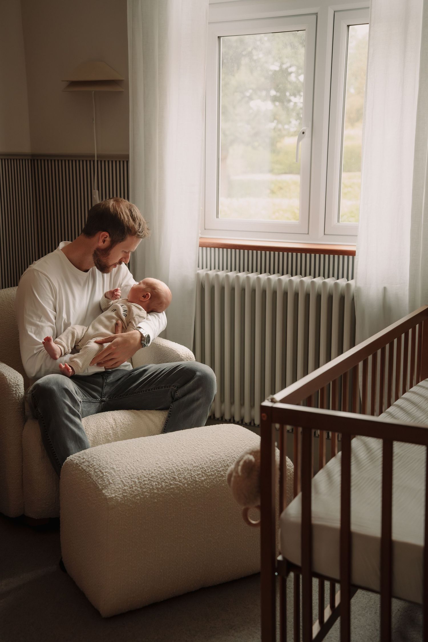 A parent in white clothing sits on a cozy cream-colored armchair next to a wooden crib in a sunlit nursery.