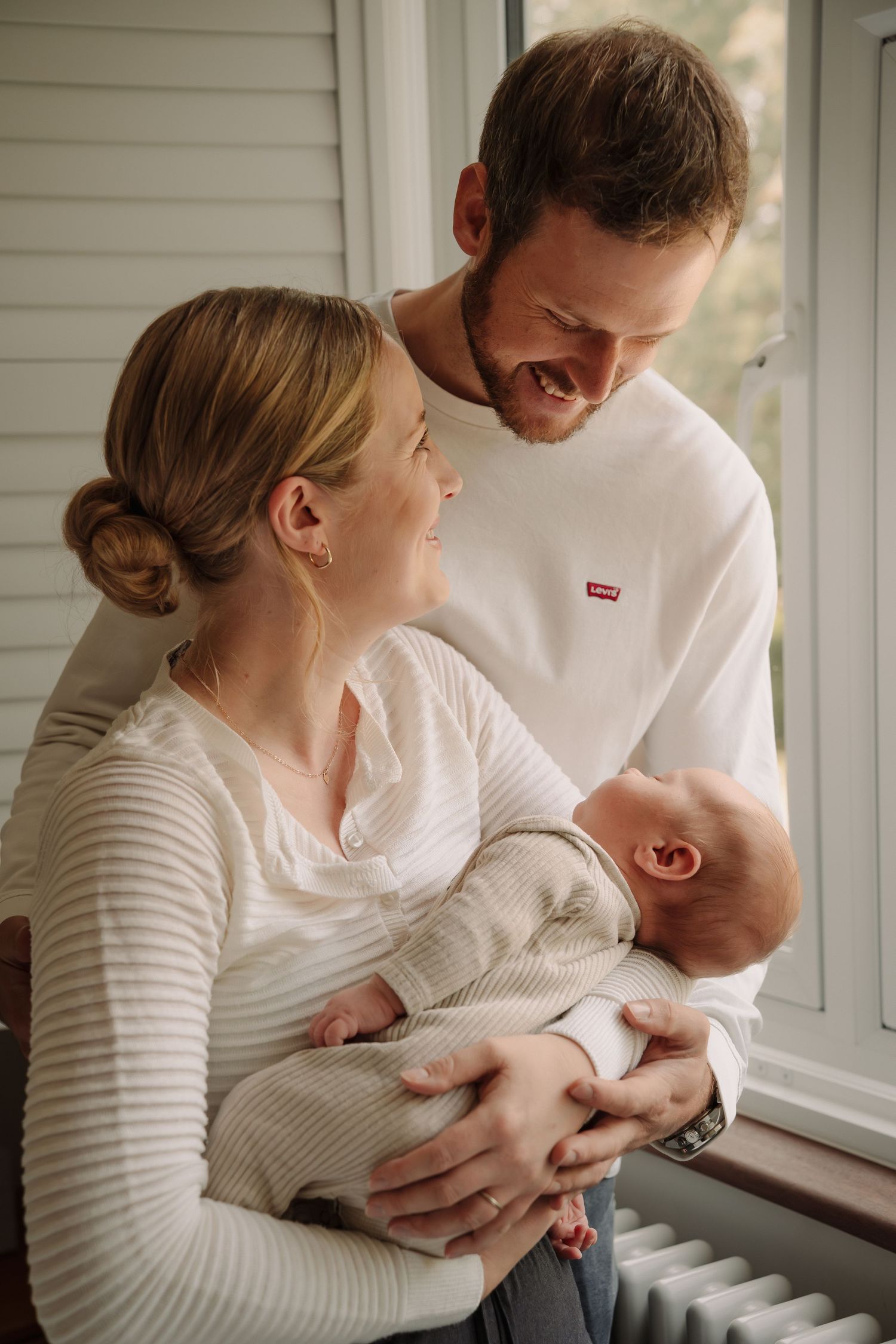 A tender family moment as parents share a loving look while holding their newborn baby near a window.