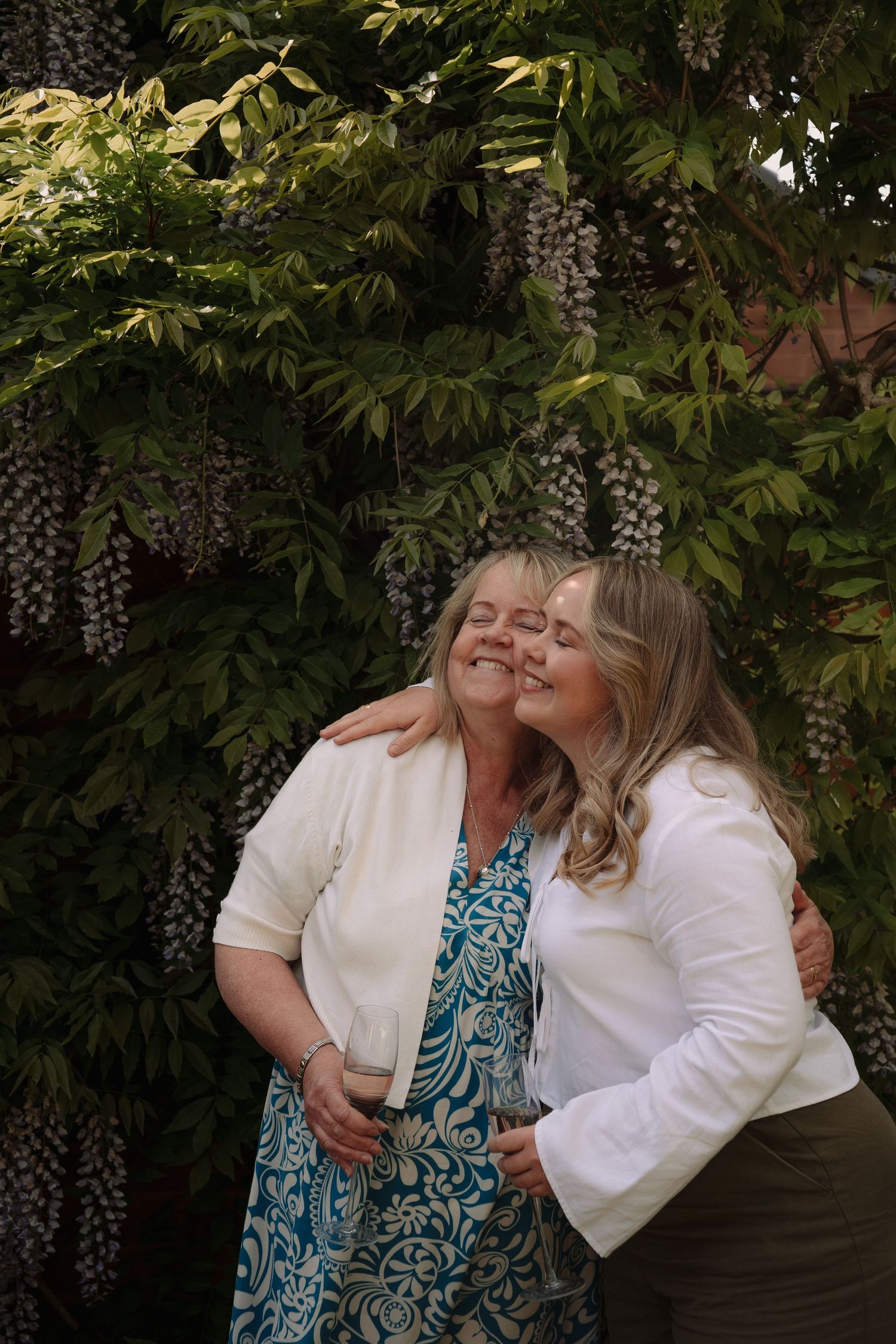 Two women embracing and smiling in front of lush wisteria blooms, wearing white jackets outdoors.