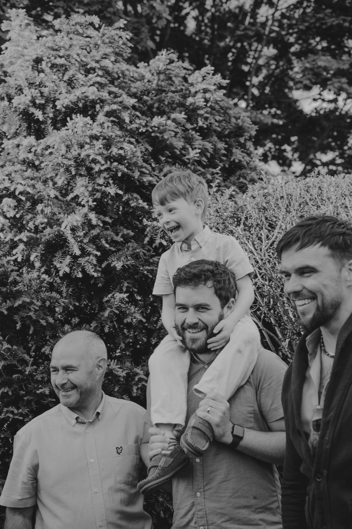 Black and white photo of three men smiling outdoors, with a laughing young boy sitting on one man's shoulders.