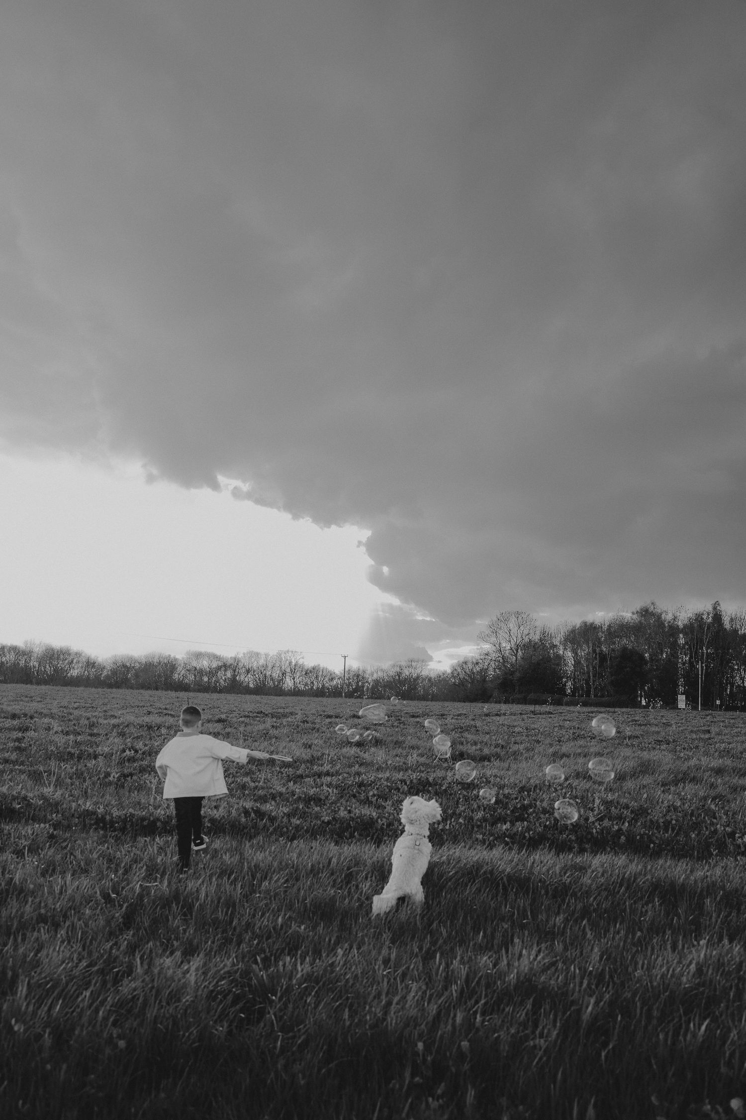 Black and white photo of a child running with a white dog in an open field under dramatic storm clouds.